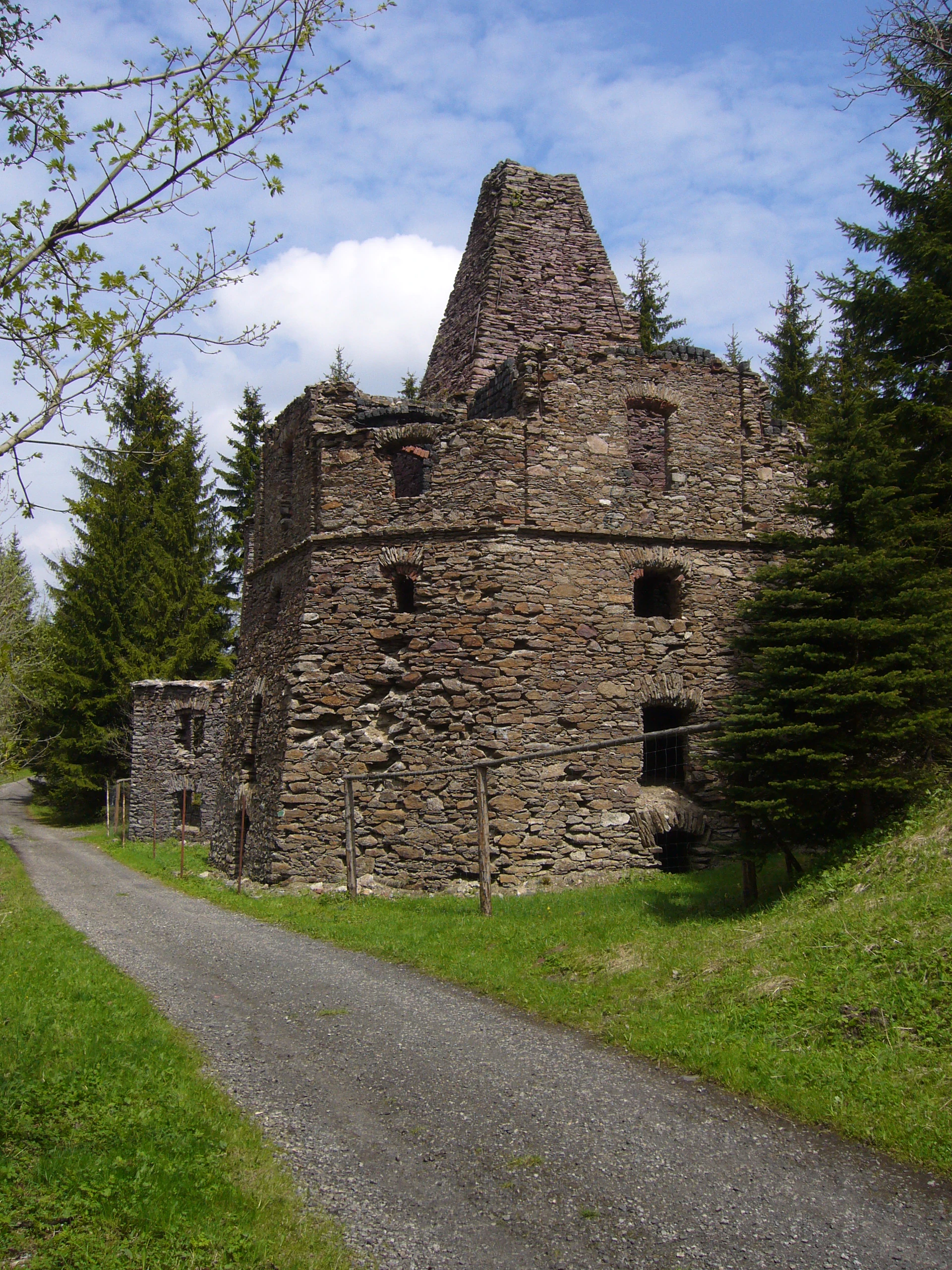 A weathered, multi-story stone lime kiln building with a tall chimney-like structure, situated next to a gravel path and surrounded by spruce trees under a partly cloudy sky.