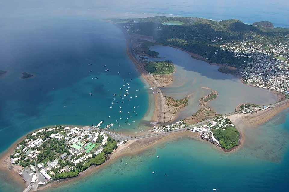 Aerial view looking down at a coastal mudflat and wetland area, bordered by a road, town structures, and the ocean with several small boats.