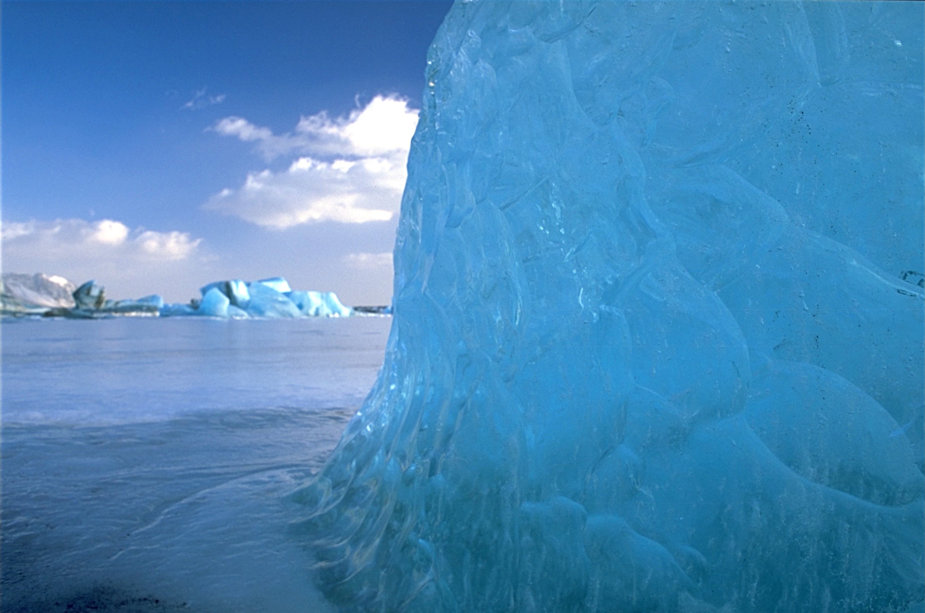Close-up of a large blue ice block on an iceberg in a glacial lagoon with distant icebergs under a clear blue sky.