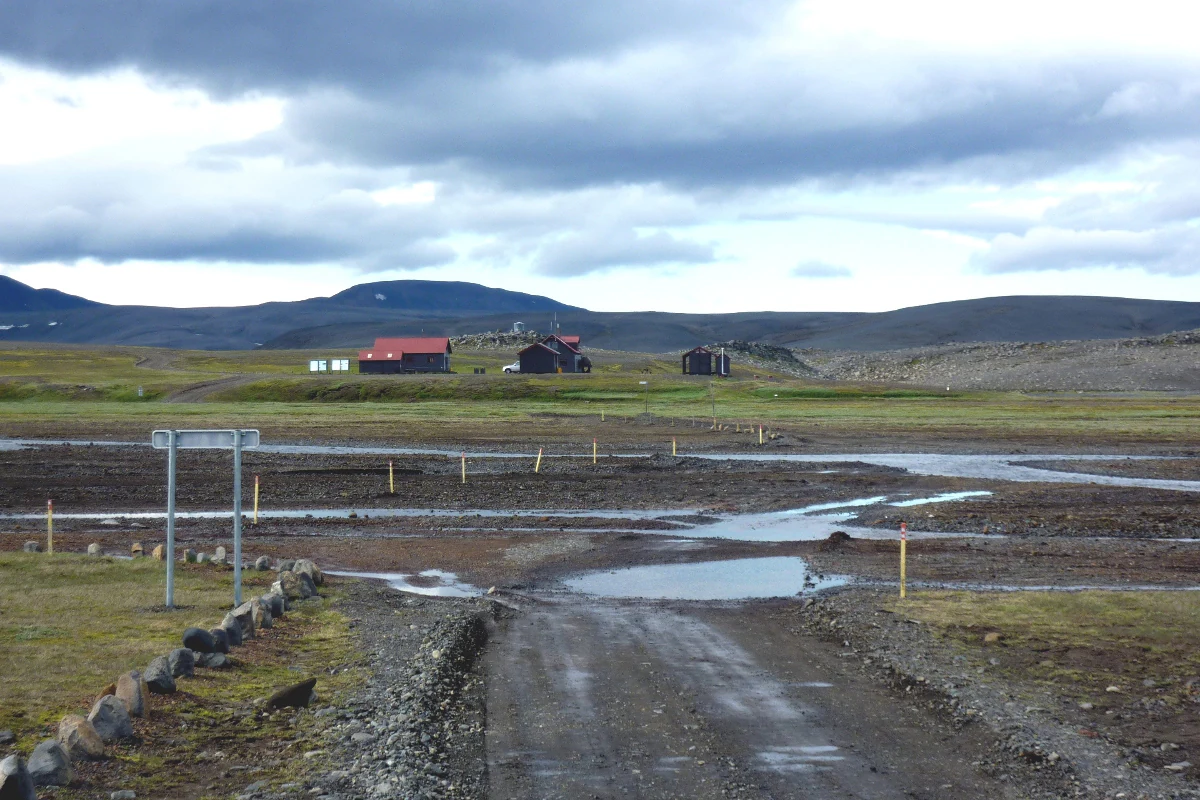 Gravel road with yellow markers, grassy fields, huts, and distant mountains under a cloudy sky