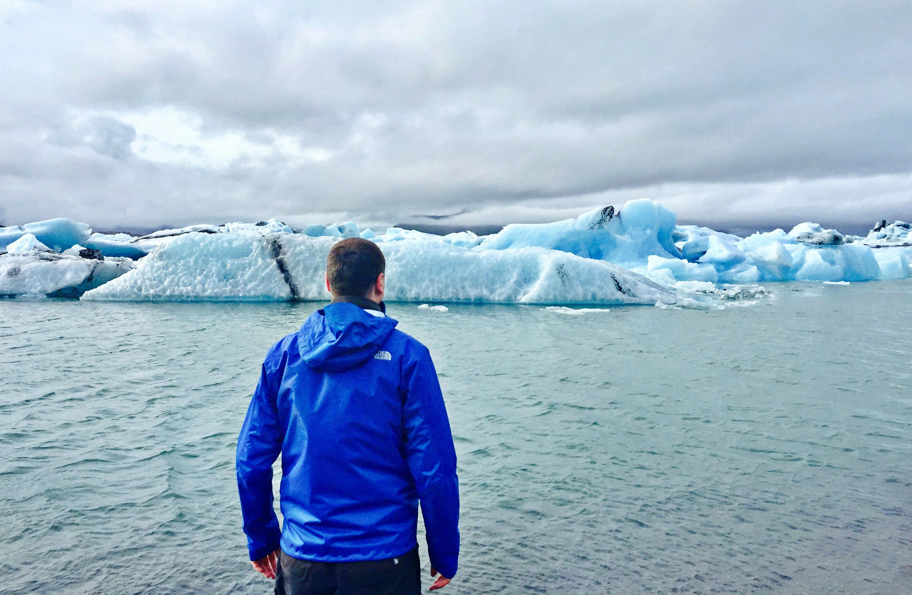 Man in blue jacket standing at the edge of a glacial lagoon with icebergs floating in the water