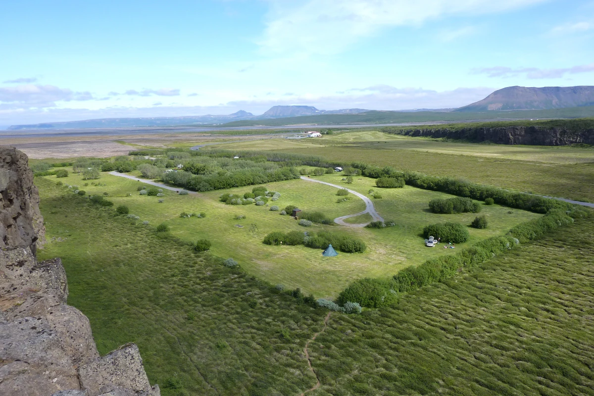 Wide landscape view of grassy fields with a winding road, scattered trees, a distant building, and a rocky cliff edge in the foreground