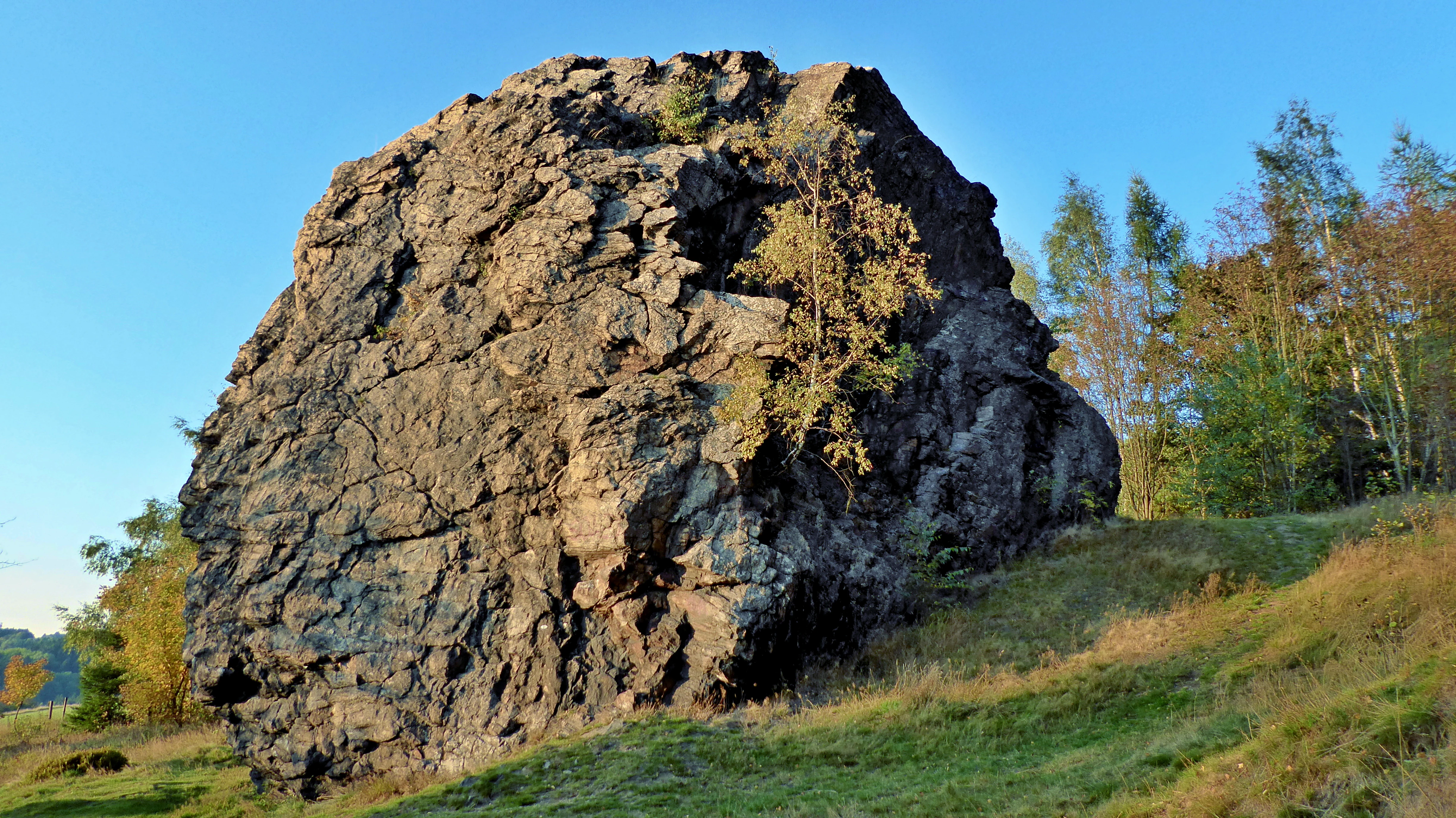 A large, weathered rock outcrop standing prominently on a grassy hillside under a clear blue sky, with trees visible in the background.