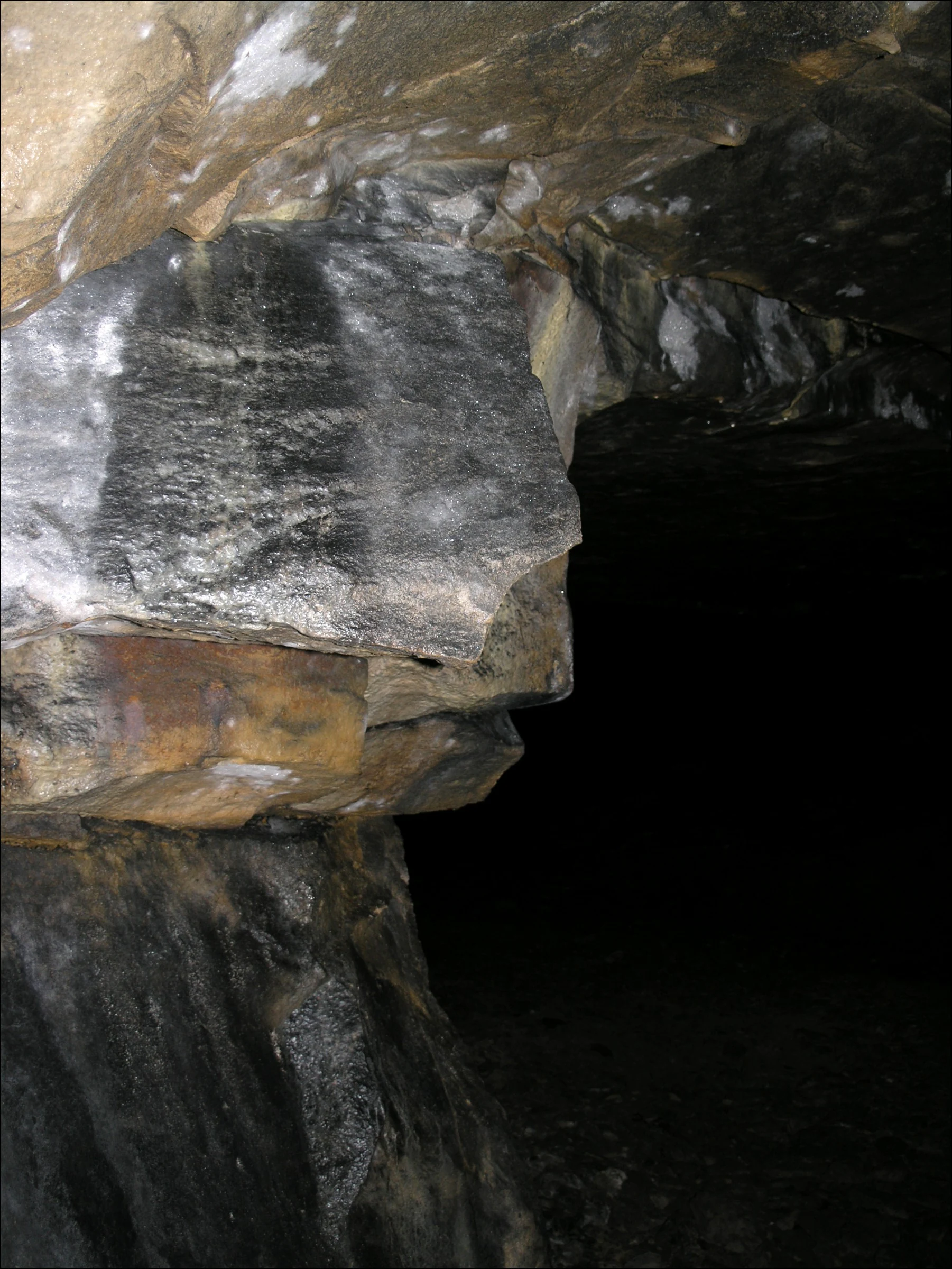 A low-angle view inside a dark underground mine or cave, showing layers of rough stone walls illuminated by artificial light against a deep shadow.
