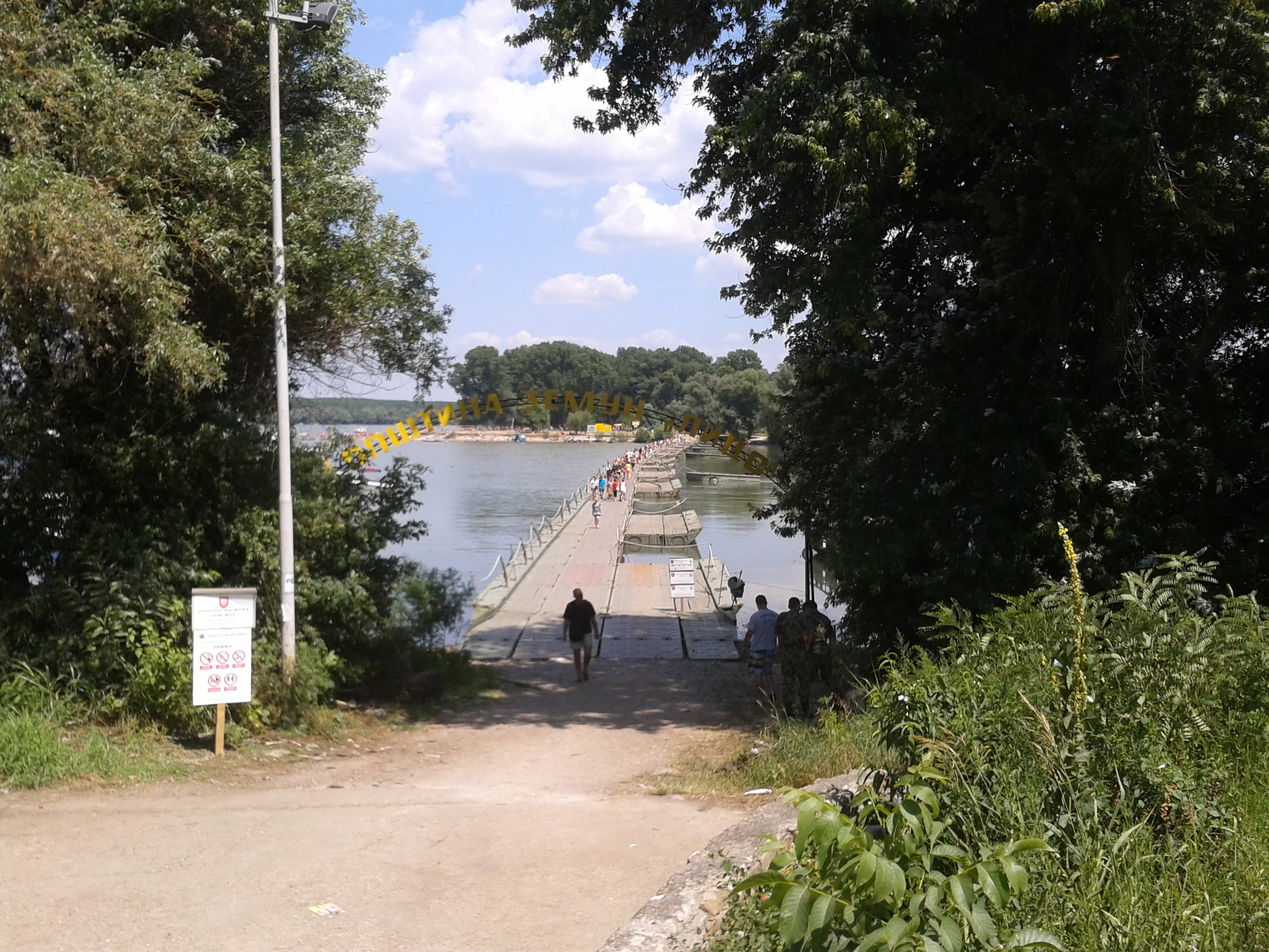 Paved pathway leading to a pontoon bridge over water, with trees on both sides and a white signpost on the left