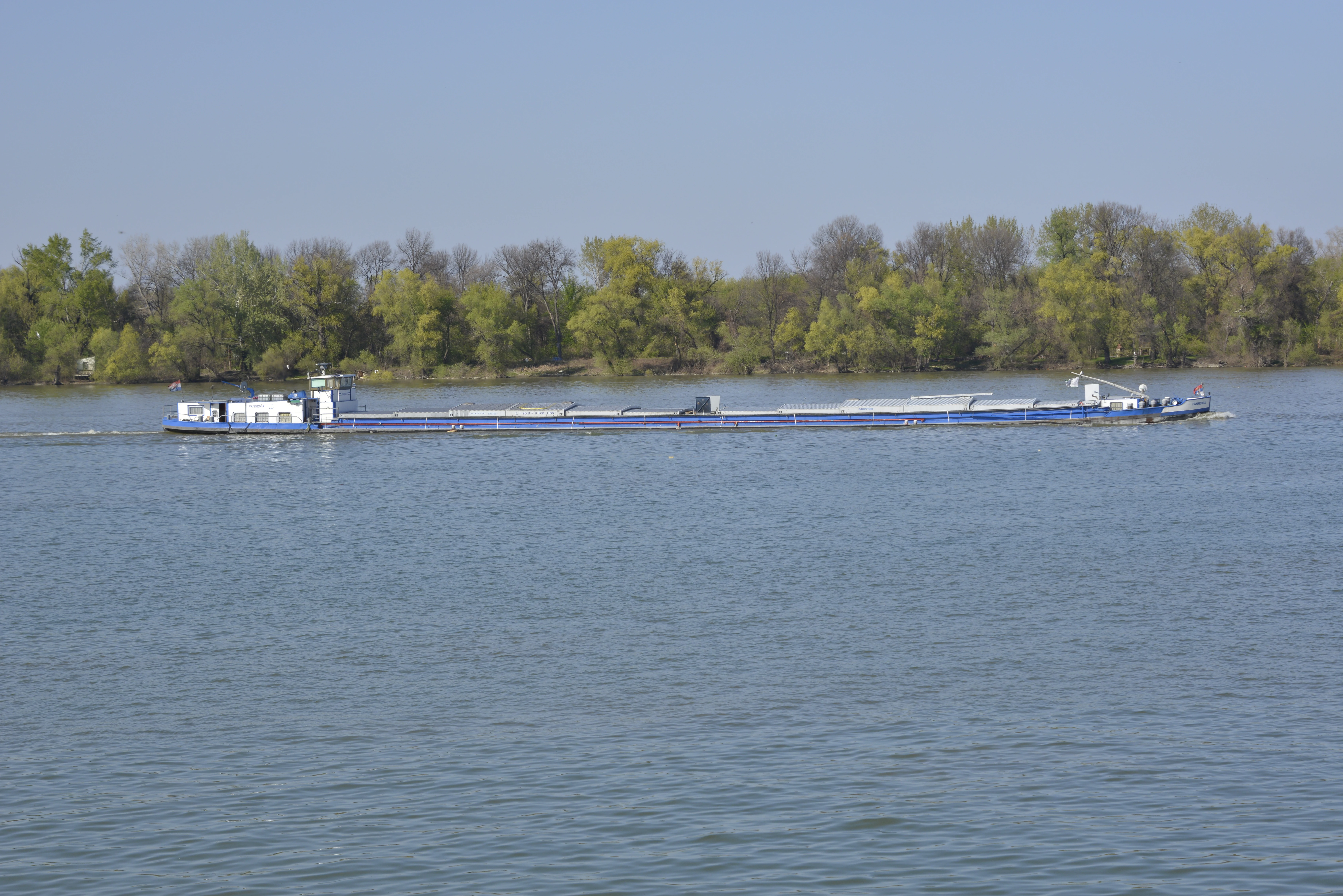Cargo barge floating on a wide river with calm waters, forested shoreline and clear blue sky