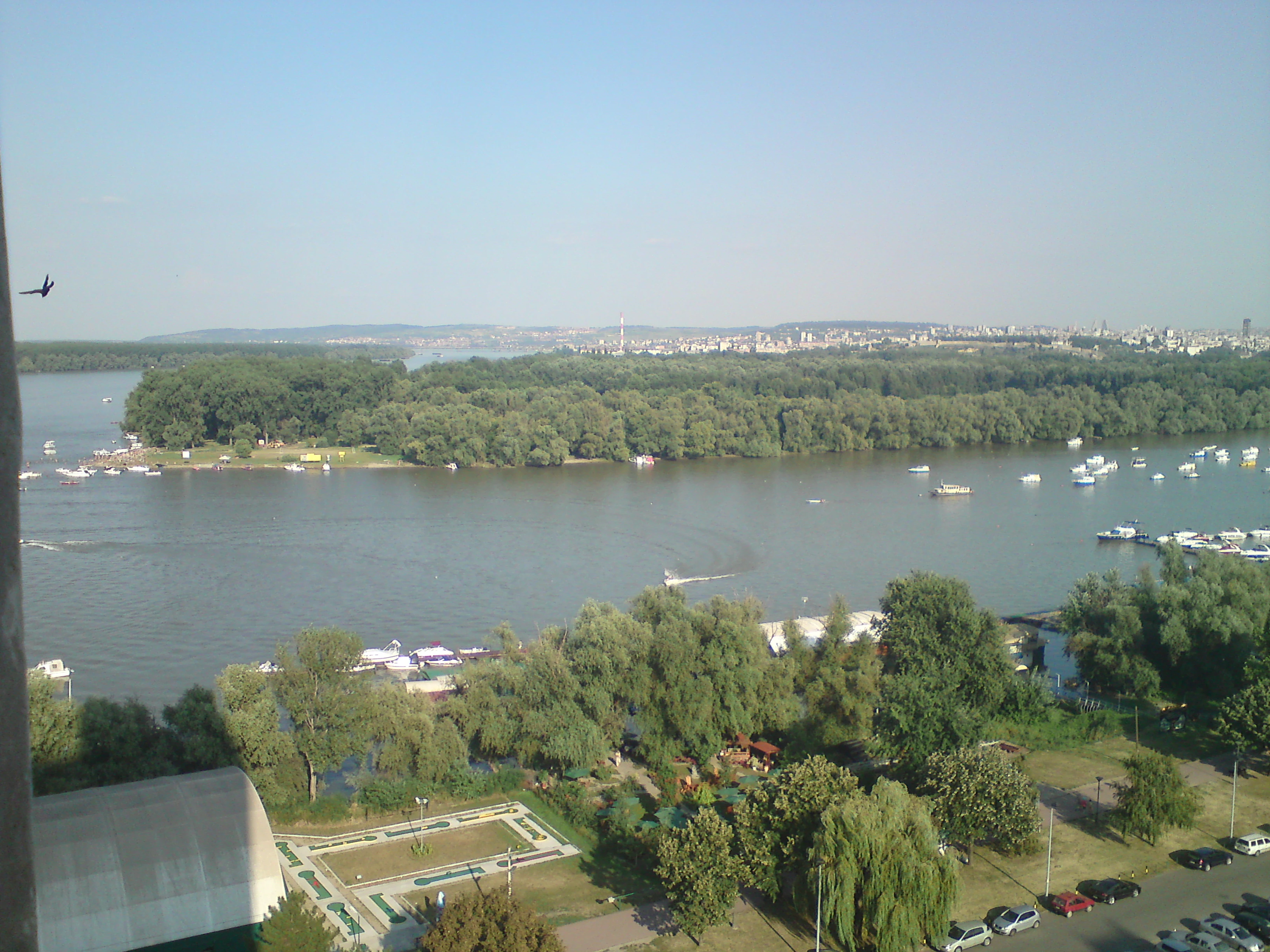 Wide view of a river with a large tree-covered island in the middle, several boats on the water, and a cityscape in the distance