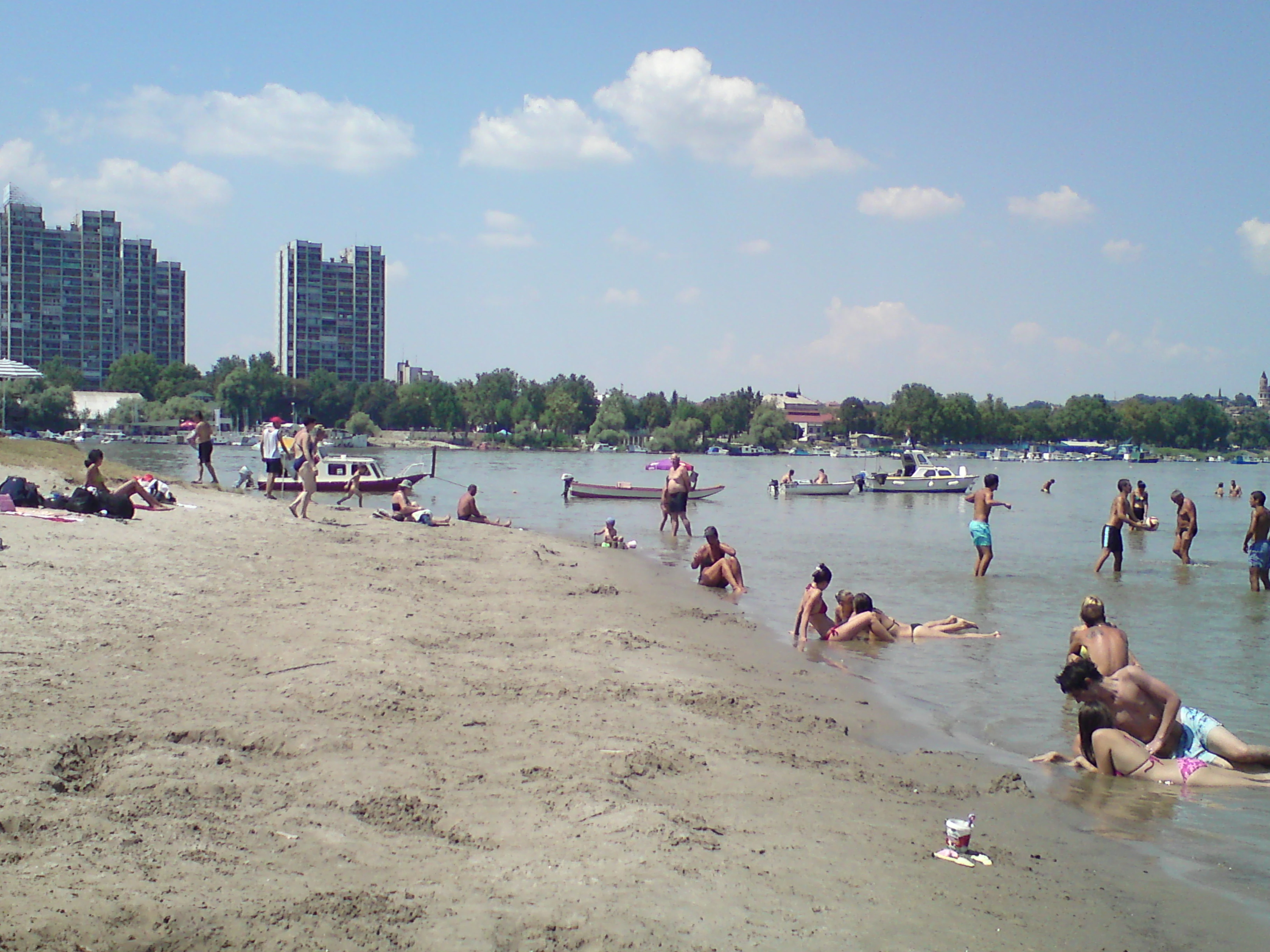 People swimming and relaxing on a sandy beach with boats on the Danube River, buildings visible in the background