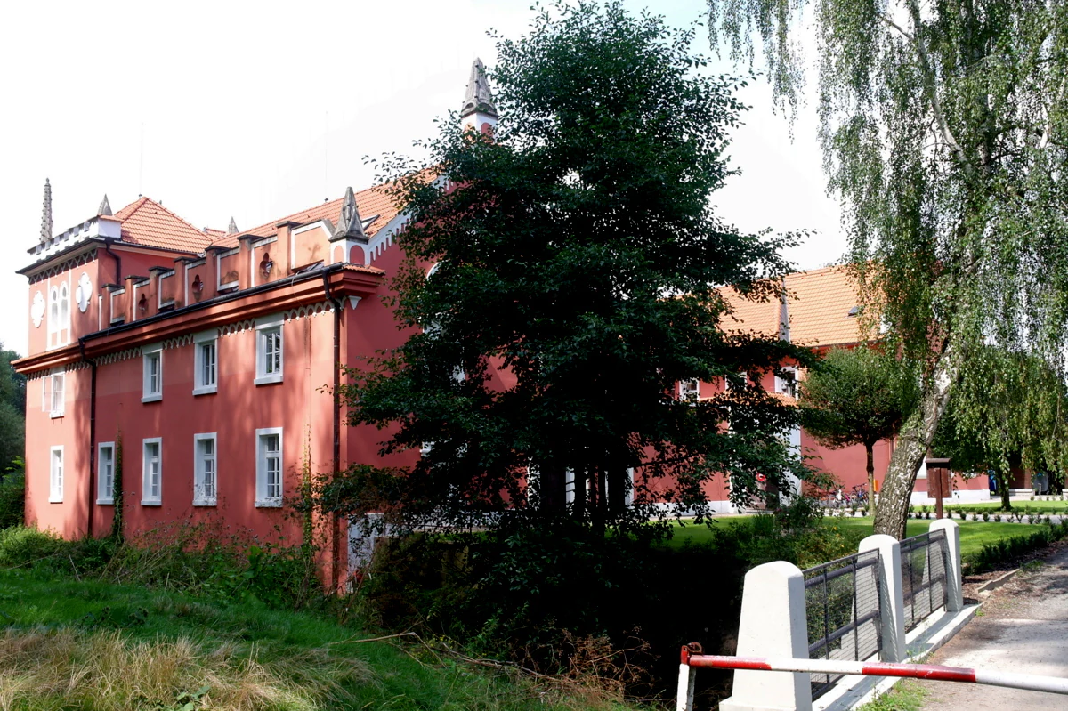 A two story red building with white window frames and a tiled roof, partially obscured by a large green tree in the foreground, situated near a small bridge.