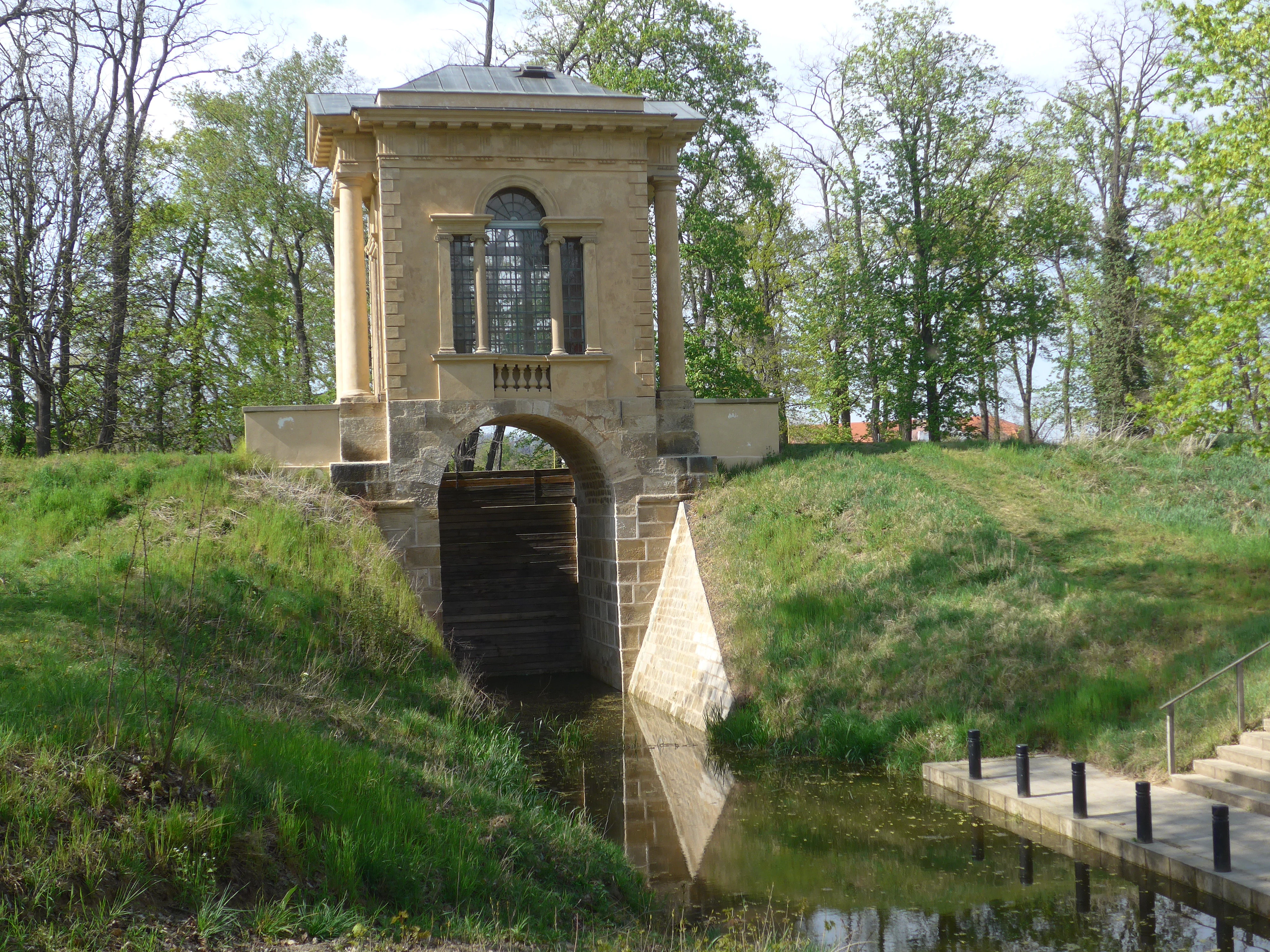 A Neoclassical stone pavilion with arched openings standing above a small stream and a stone boat landing in a wooded park setting.