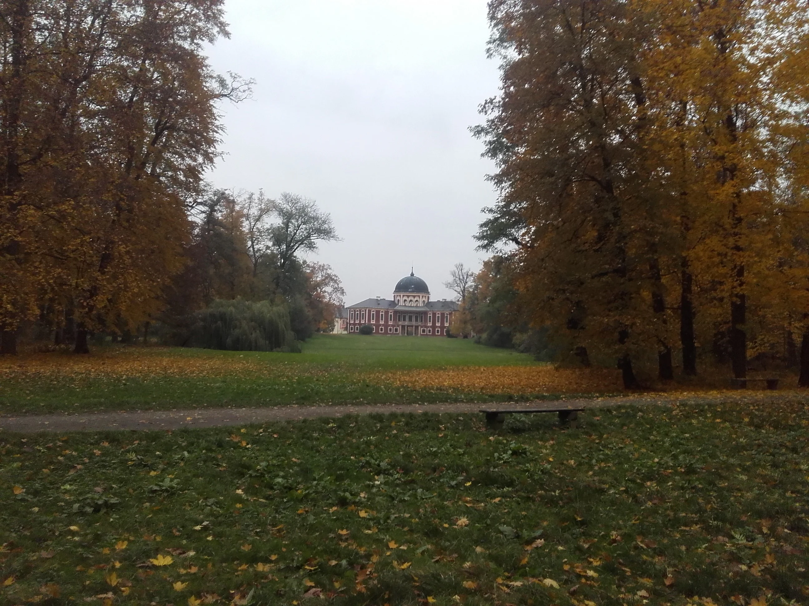 A wide angle view of the Veltrusy Mansion situated at the end of a long lawn, surrounded by autumn trees with yellow and orange leaves.