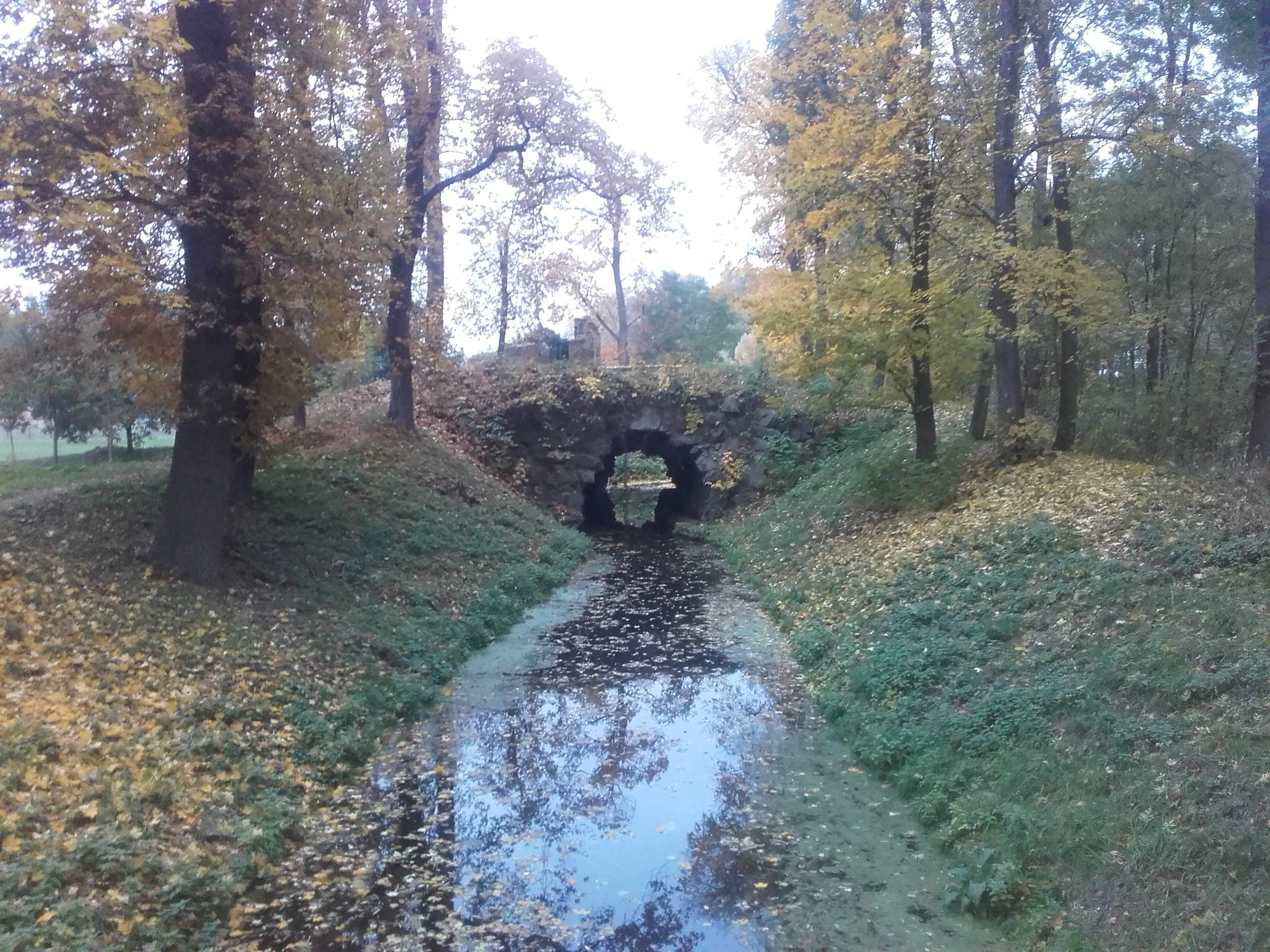 An old, small stone archway or ruin built into a grassy embankment, surrounded by trees with autumn foliage, spanning over a small, stagnant stream.