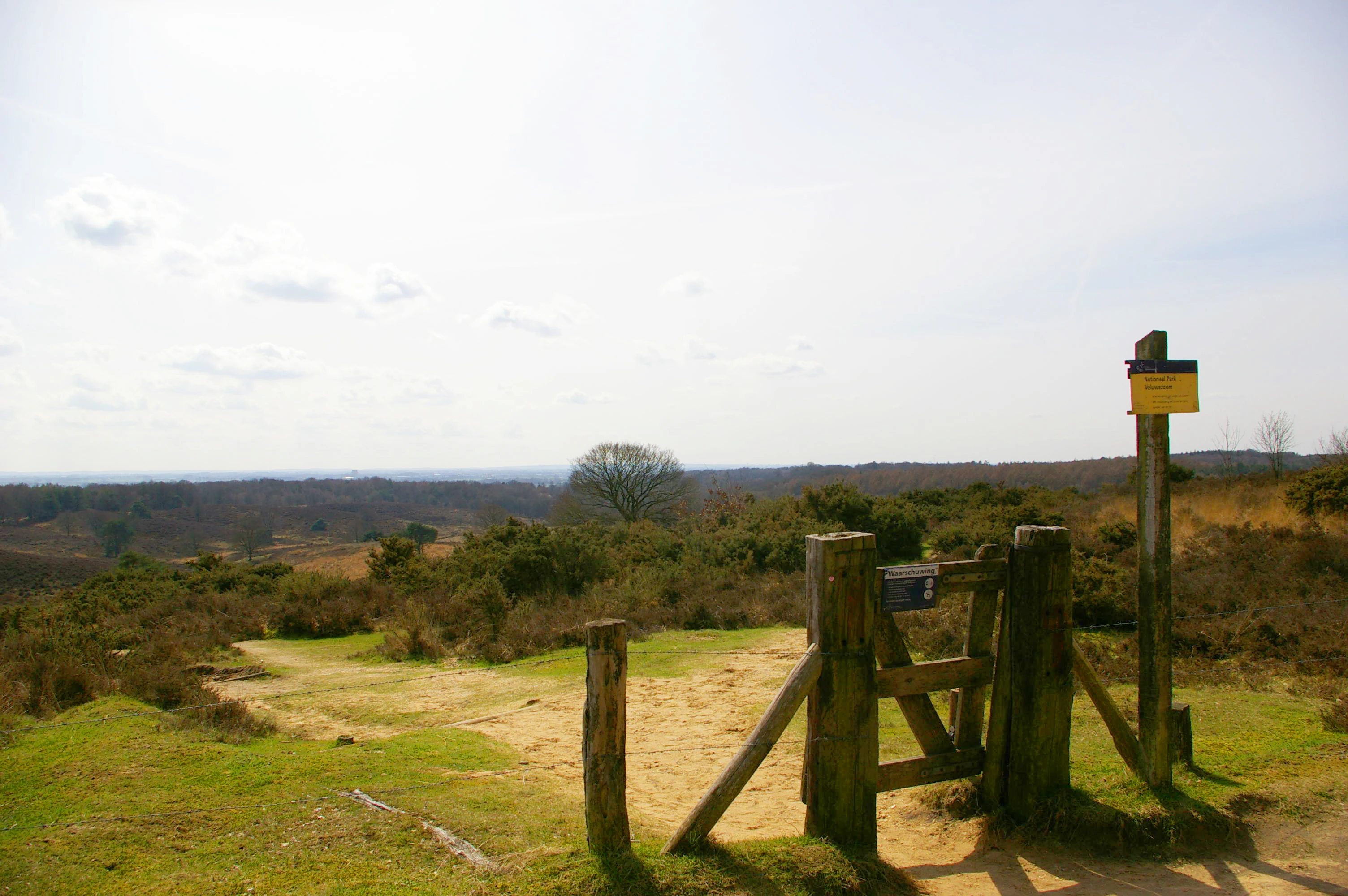 Wooden gate structure with yellow signpost, grassy field, and distant hilly landscape under partly cloudy sky