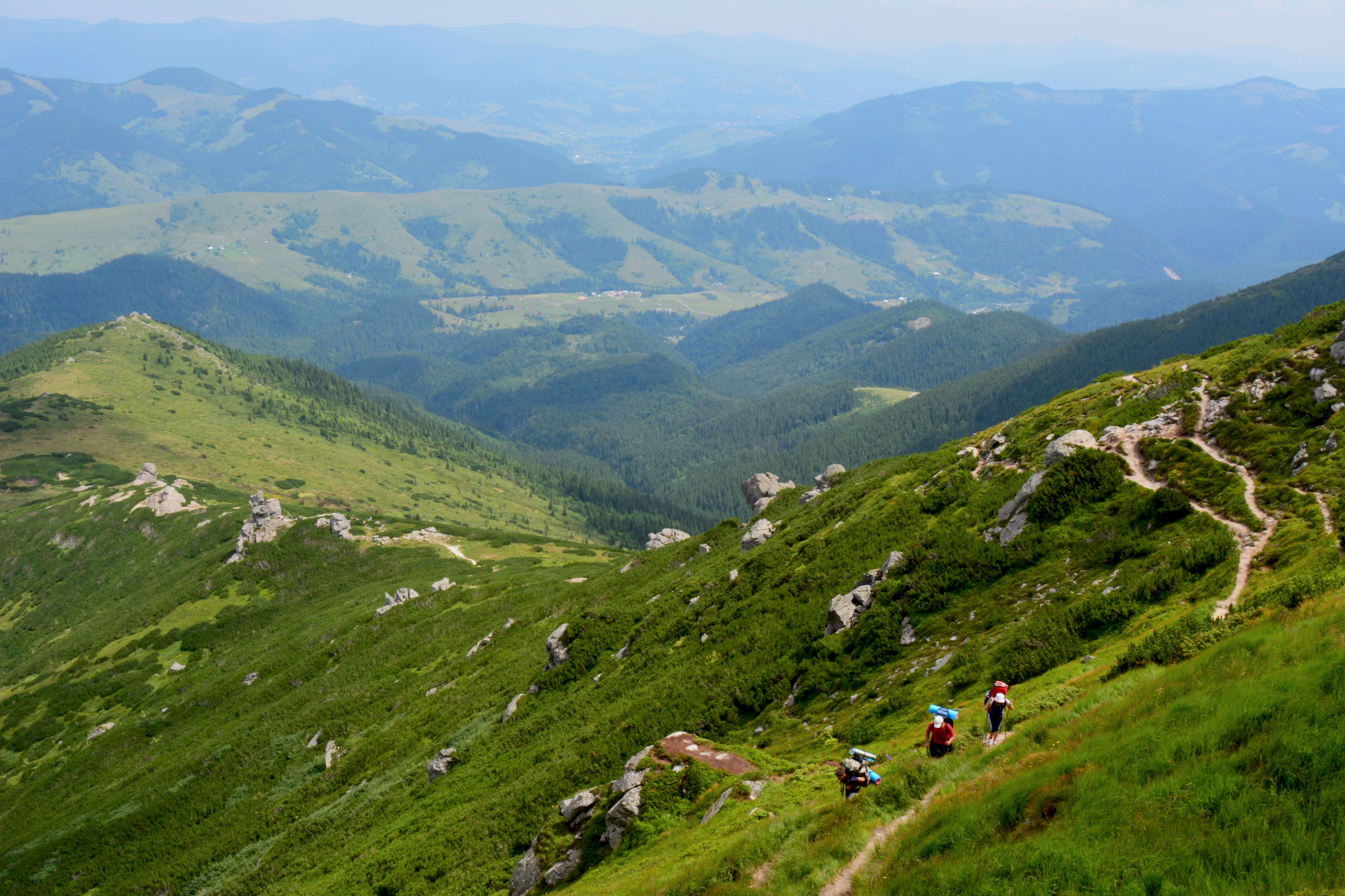 Mountain trail with hikers on a grassy slope, surrounded by rolling hills and valleys