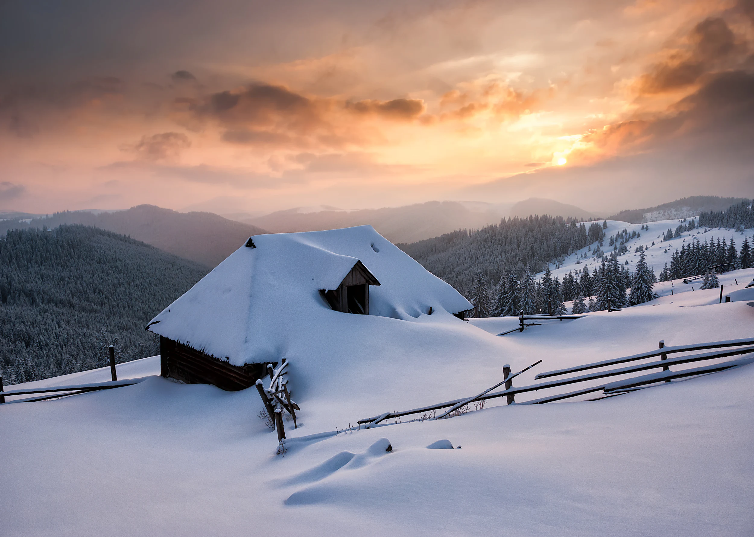 Snow-covered wooden cabin with a sloped roof, surrounded by a snow-covered landscape, wooden fence, and mountainous terrain under a partly cloudy sky with sunset hues