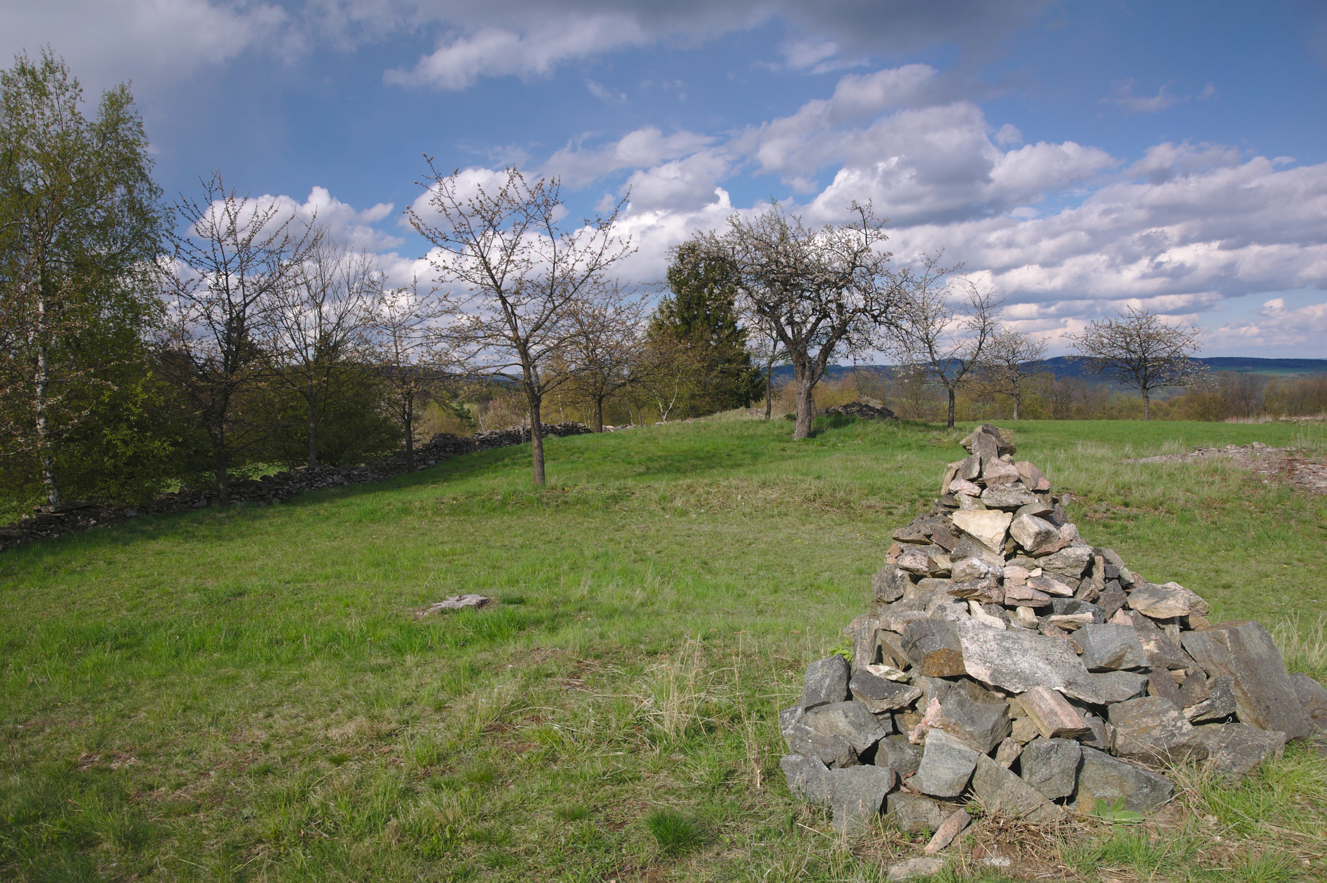 A green hillside with a stone cairn in the foreground, scattered deciduous trees, and a distant treeline under a partly cloudy blue sky.