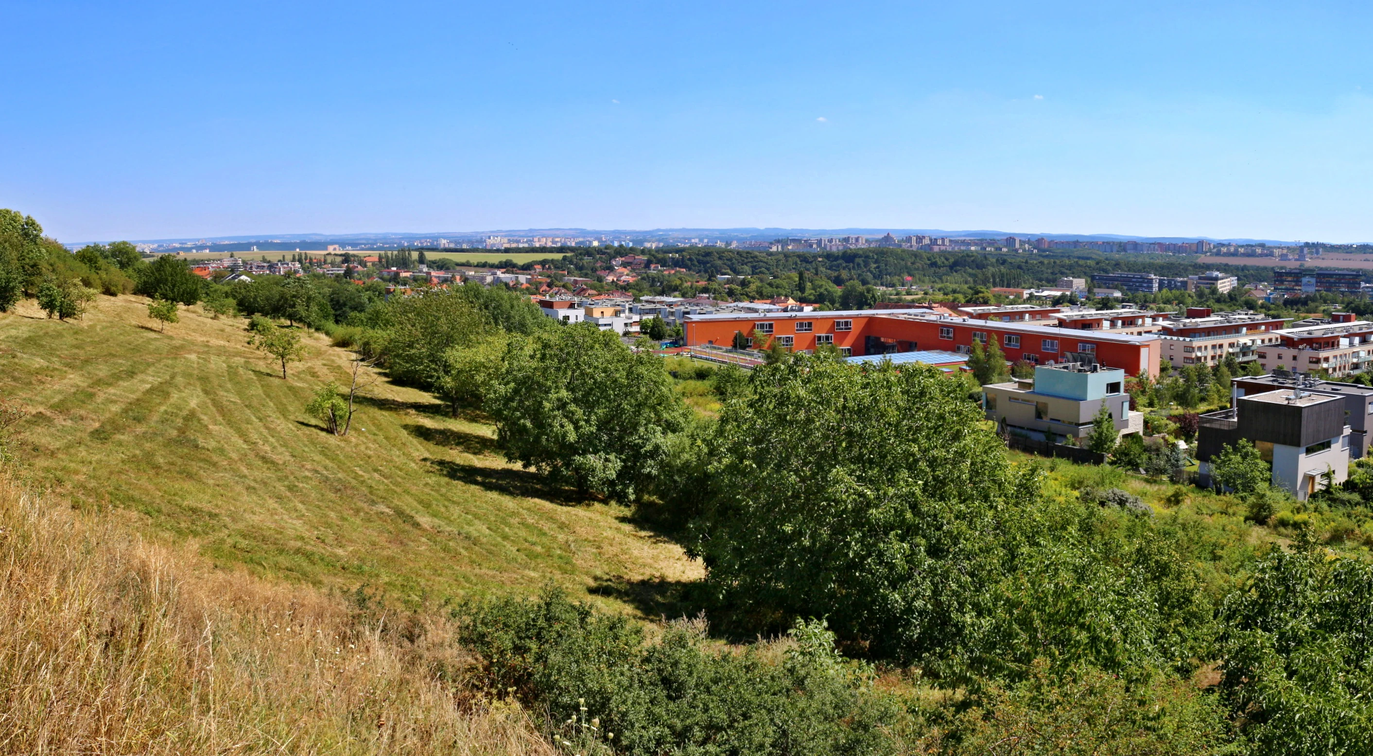 A wide angle view from the grassy, sloped edge of the Vidoule plateau looking down toward a modern school complex and residential area in Prague.