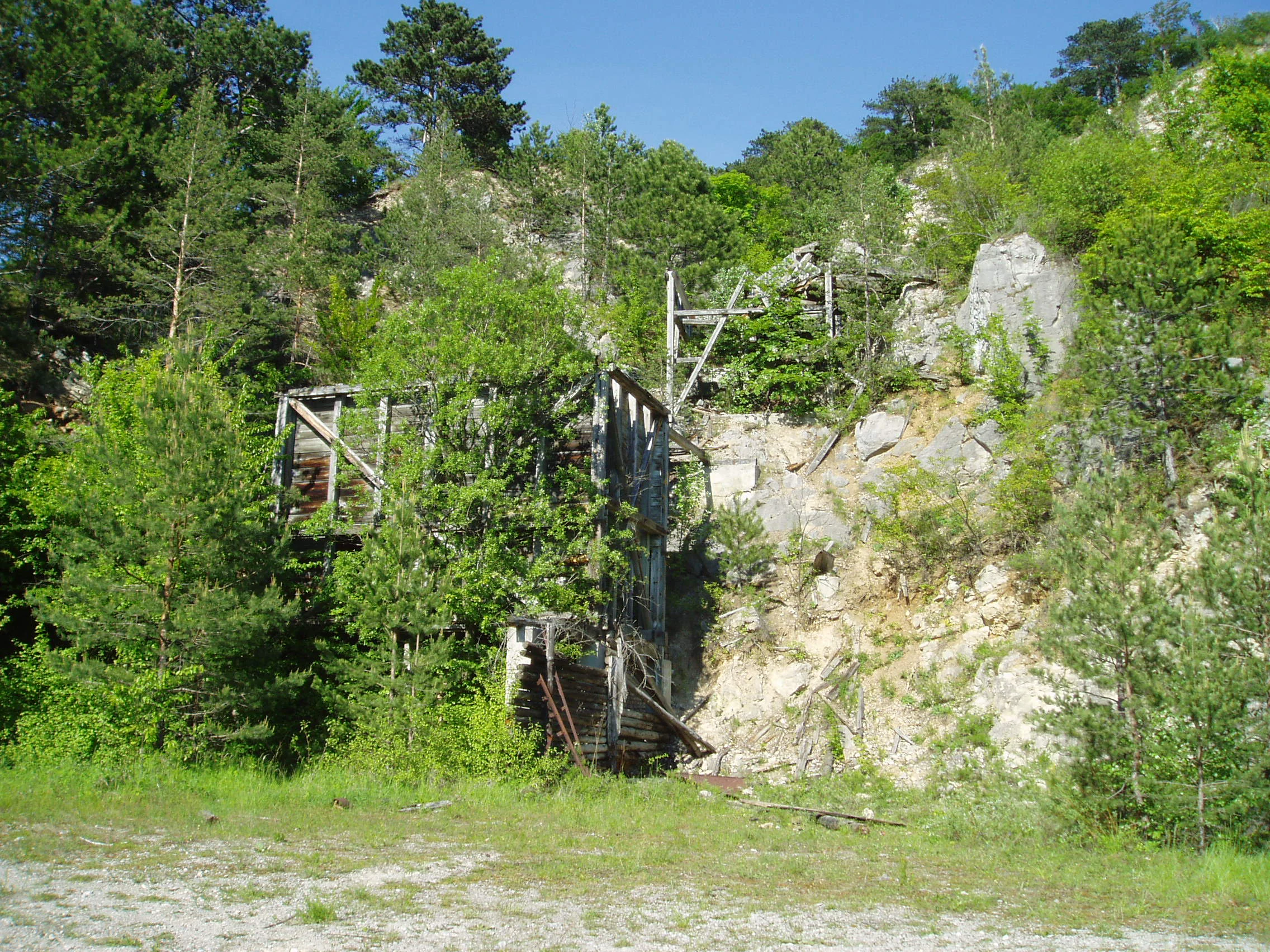 An overgrown, abandoned wooden mine structure set against a rocky limestone cliff surrounded by dense green forest.