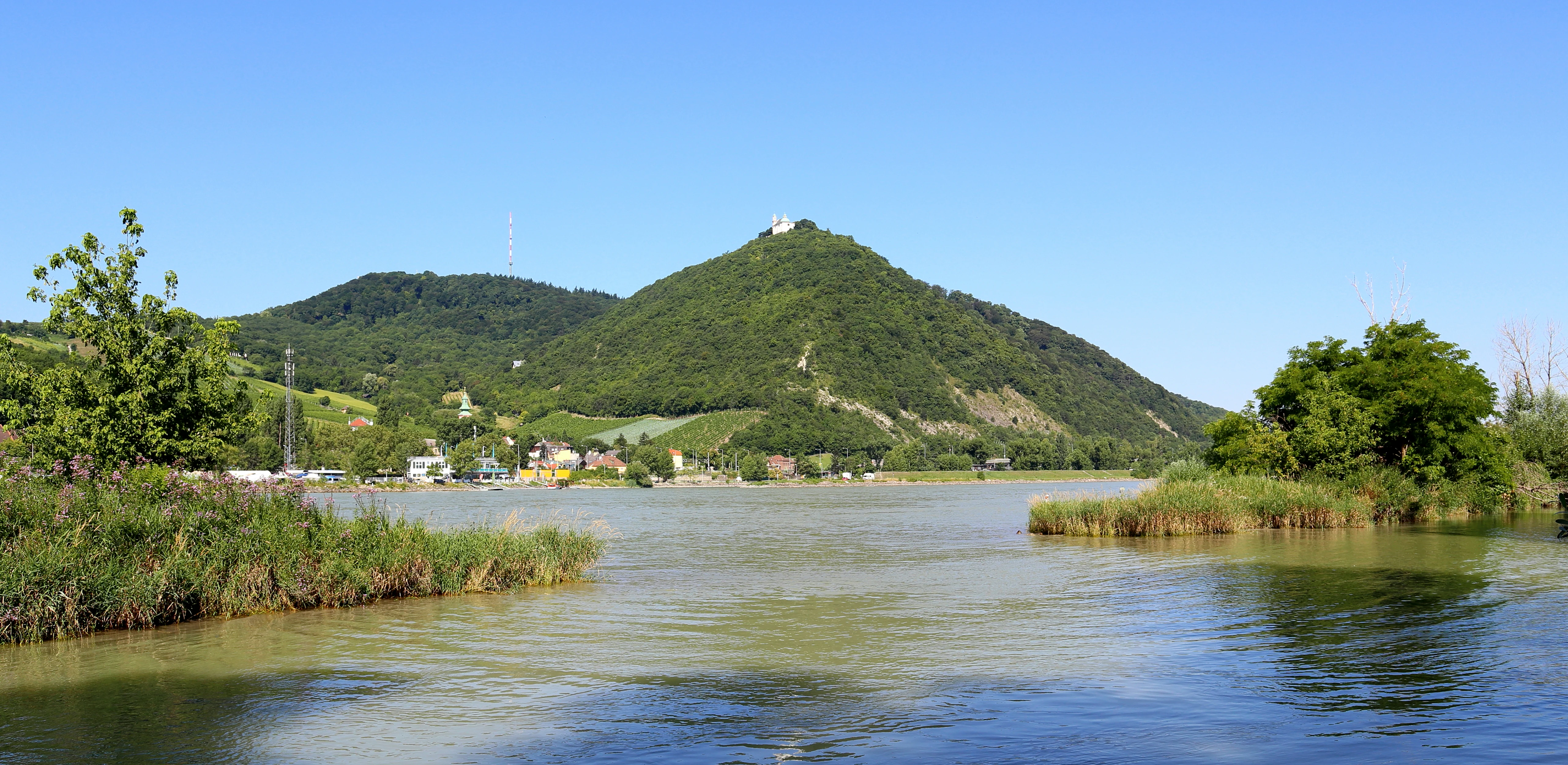 A wide, eye-level shot of a calm river in the foreground, with lush, tree-covered hills rising in the background under a clear blue sky; a small church is visible on the summit of the central peak.