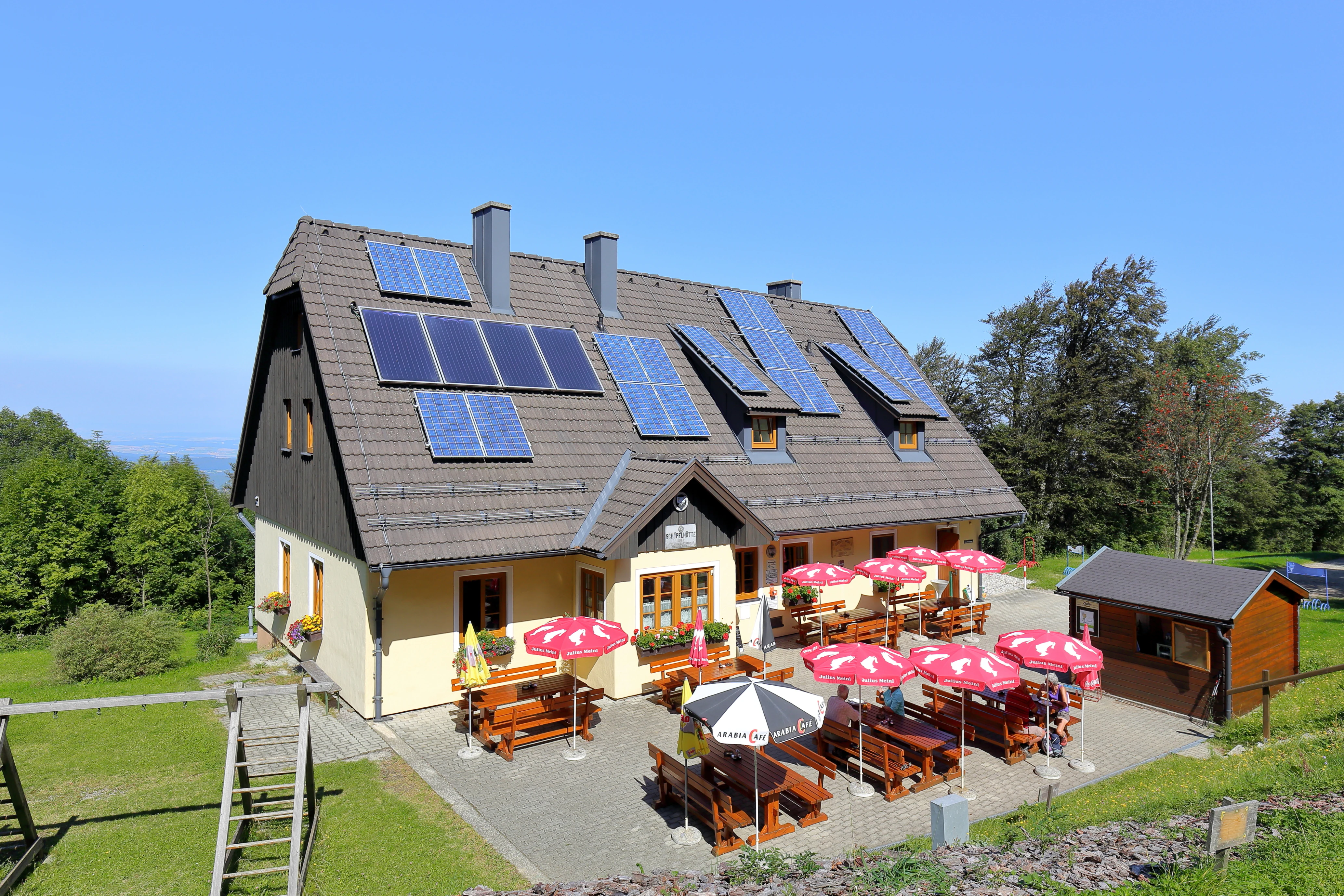 A mountain refuge building with solar panels on its roof, surrounded by a terrace with red umbrellas and wooden tables in the Vienna Woods.