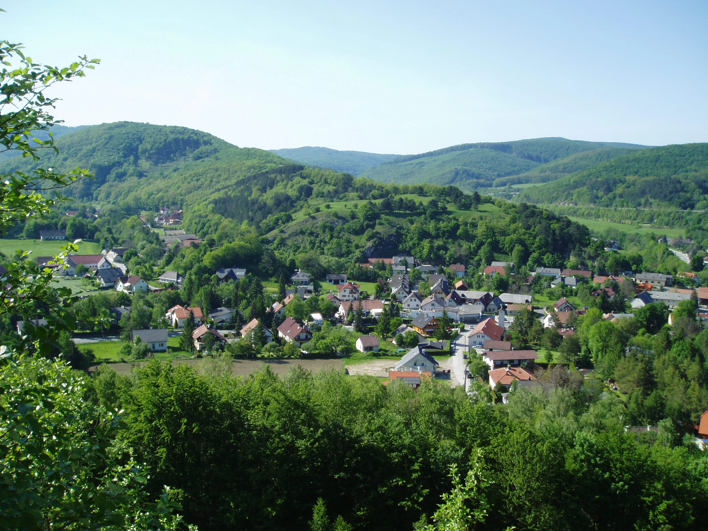 A high-angle scenic view of the village of Alland in Austria, showing houses surrounded by lush green deciduous forests and rolling hills under a clear sky.