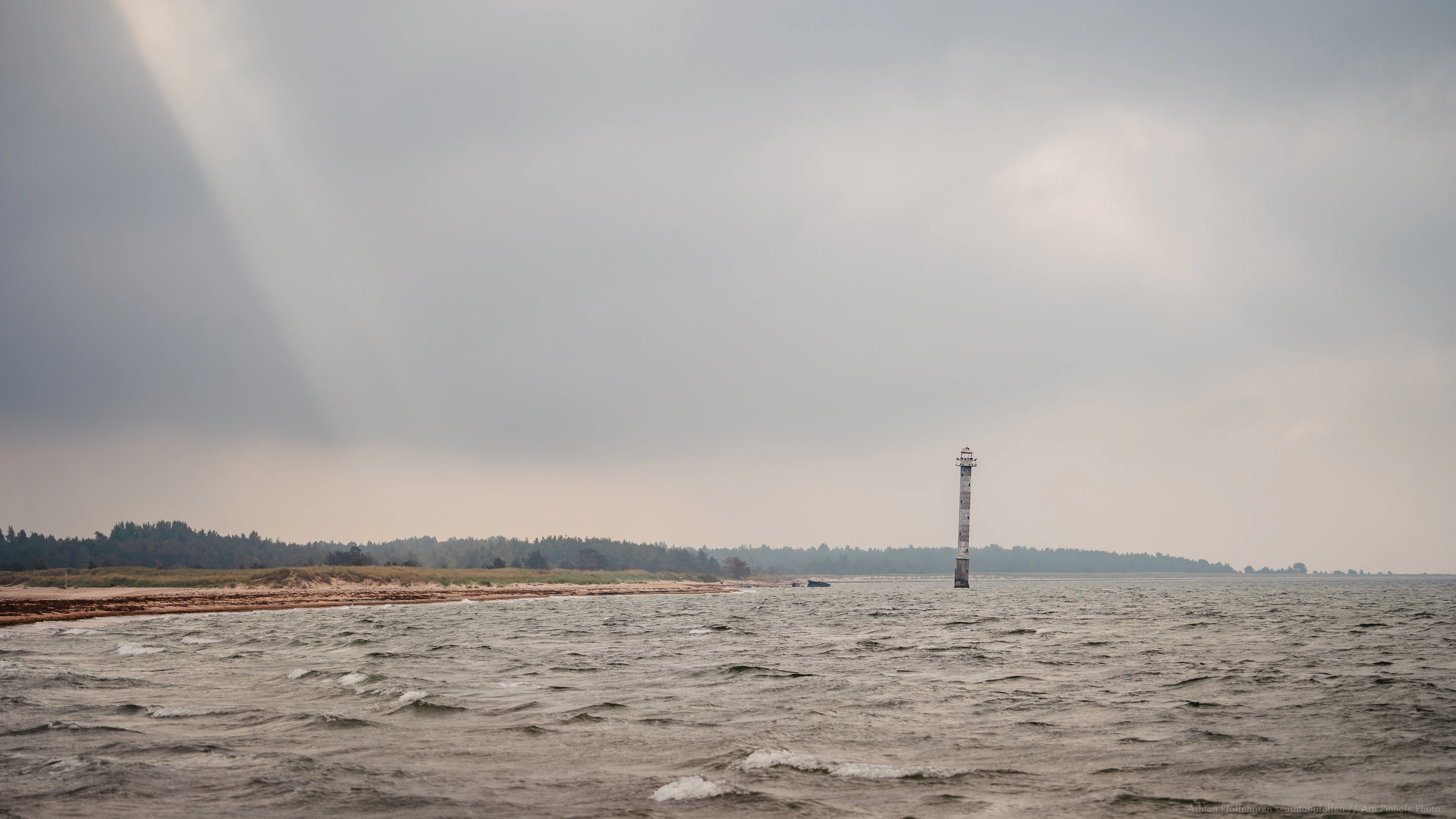 Choppy sea with distant lighthouse under overcast sky, shoreline with trees in background
