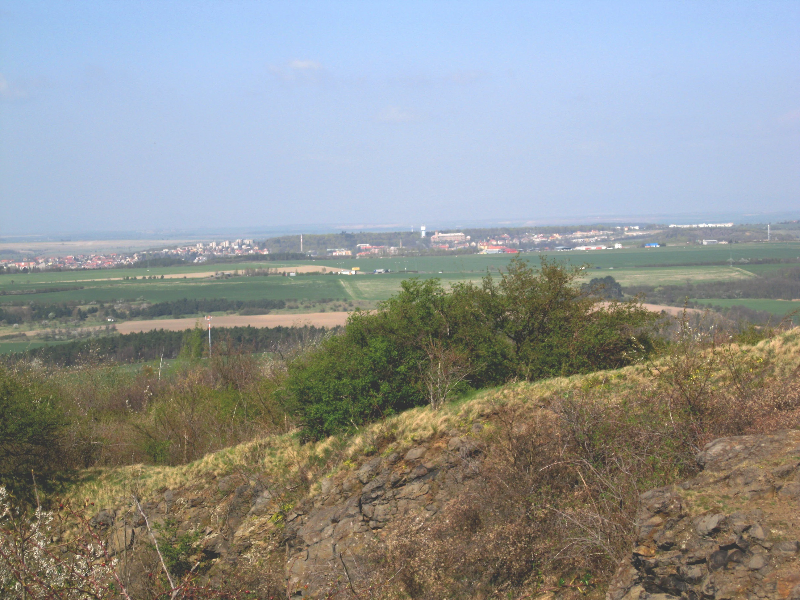 A distant view looking down from a grassy, rocky hillside across a rural landscape towards a distant settlement under a clear sky.