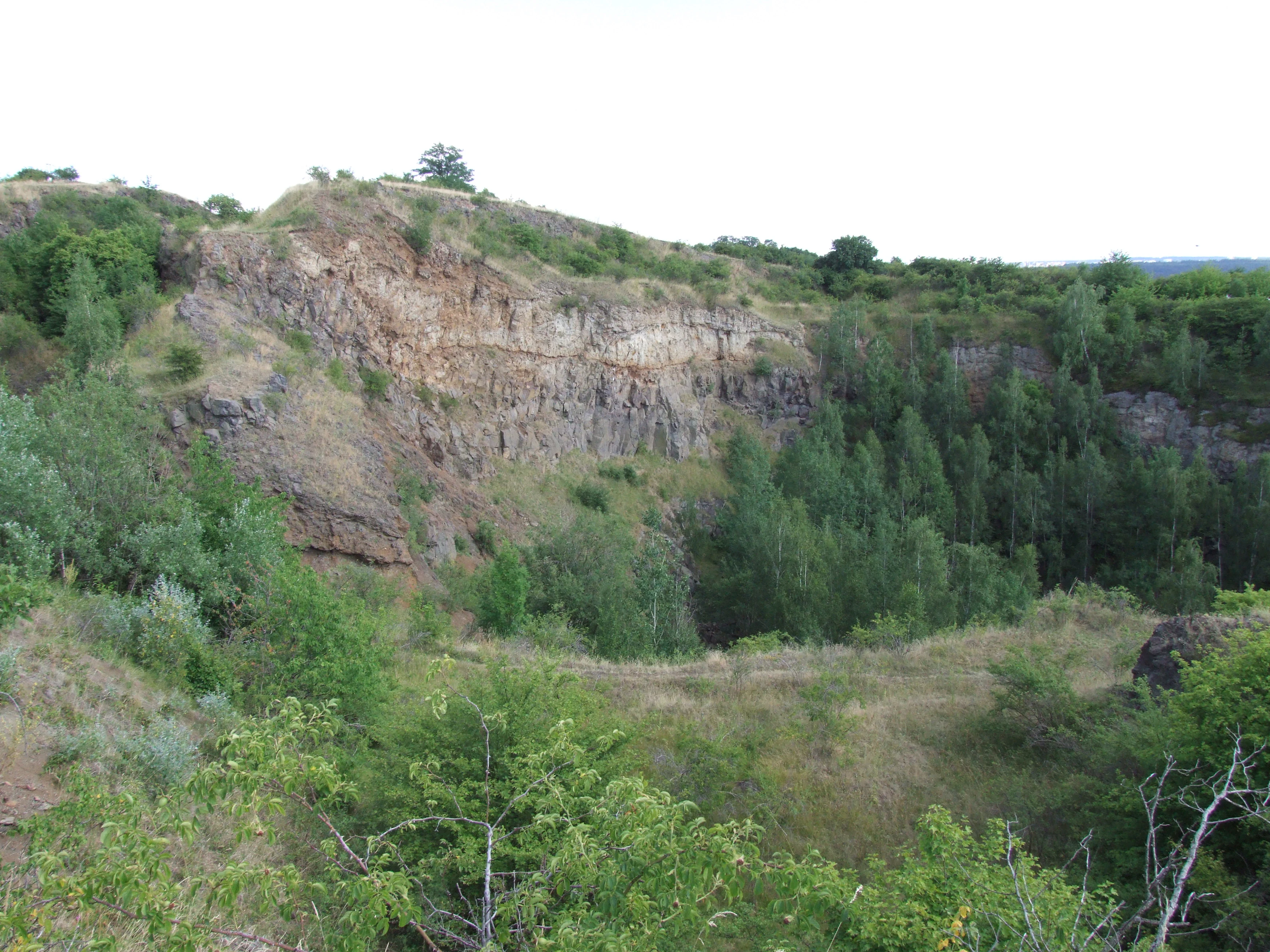 A wide view of an abandoned quarry at Vinařická hora, featuring steep, rocky cliffs and slopes covered in patches of green trees and tall grass.