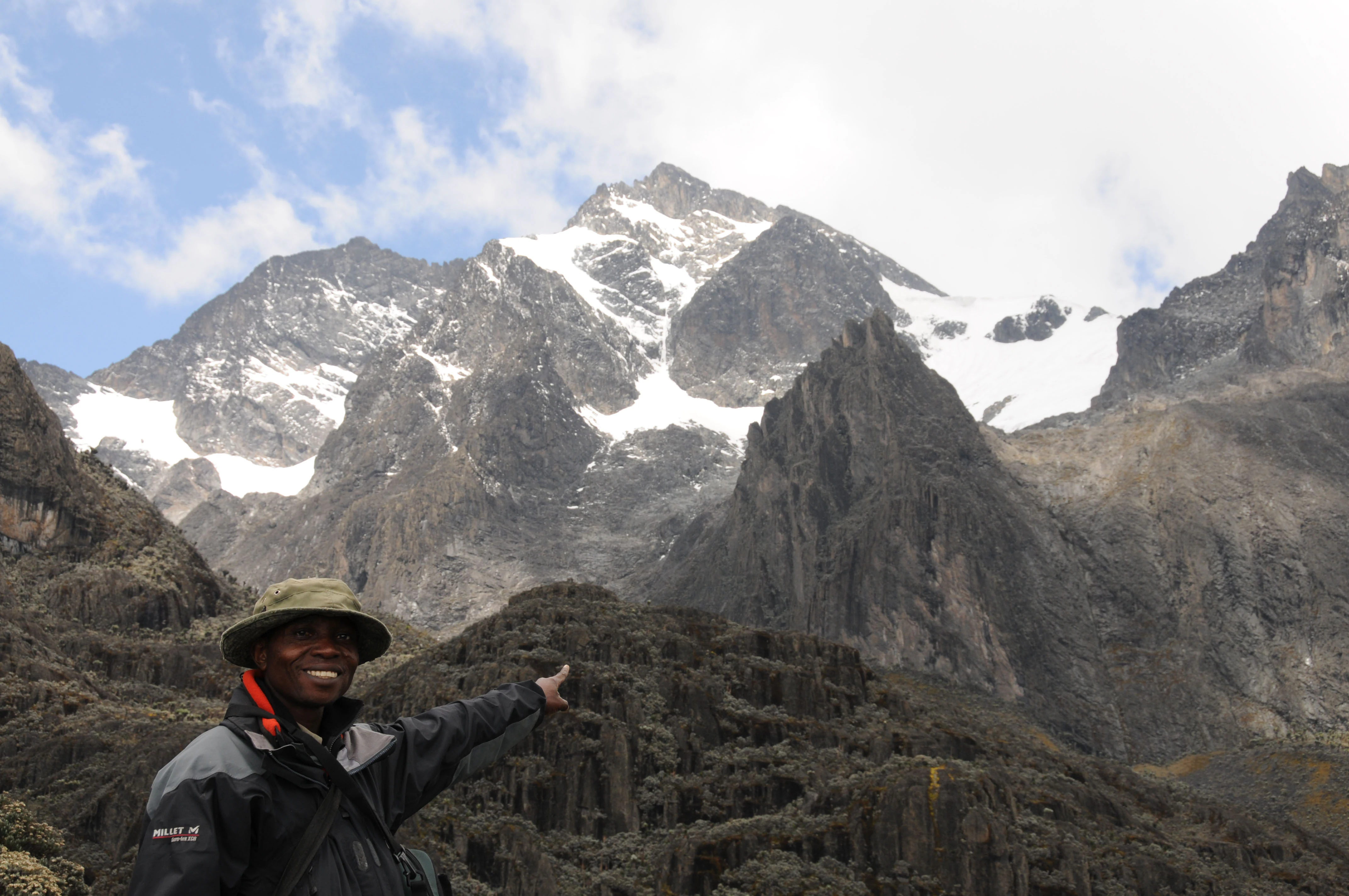 A person in a green hat and dark jacket stands in front of rugged, snow-capped mountains with rocky terrain under a partly cloudy sky