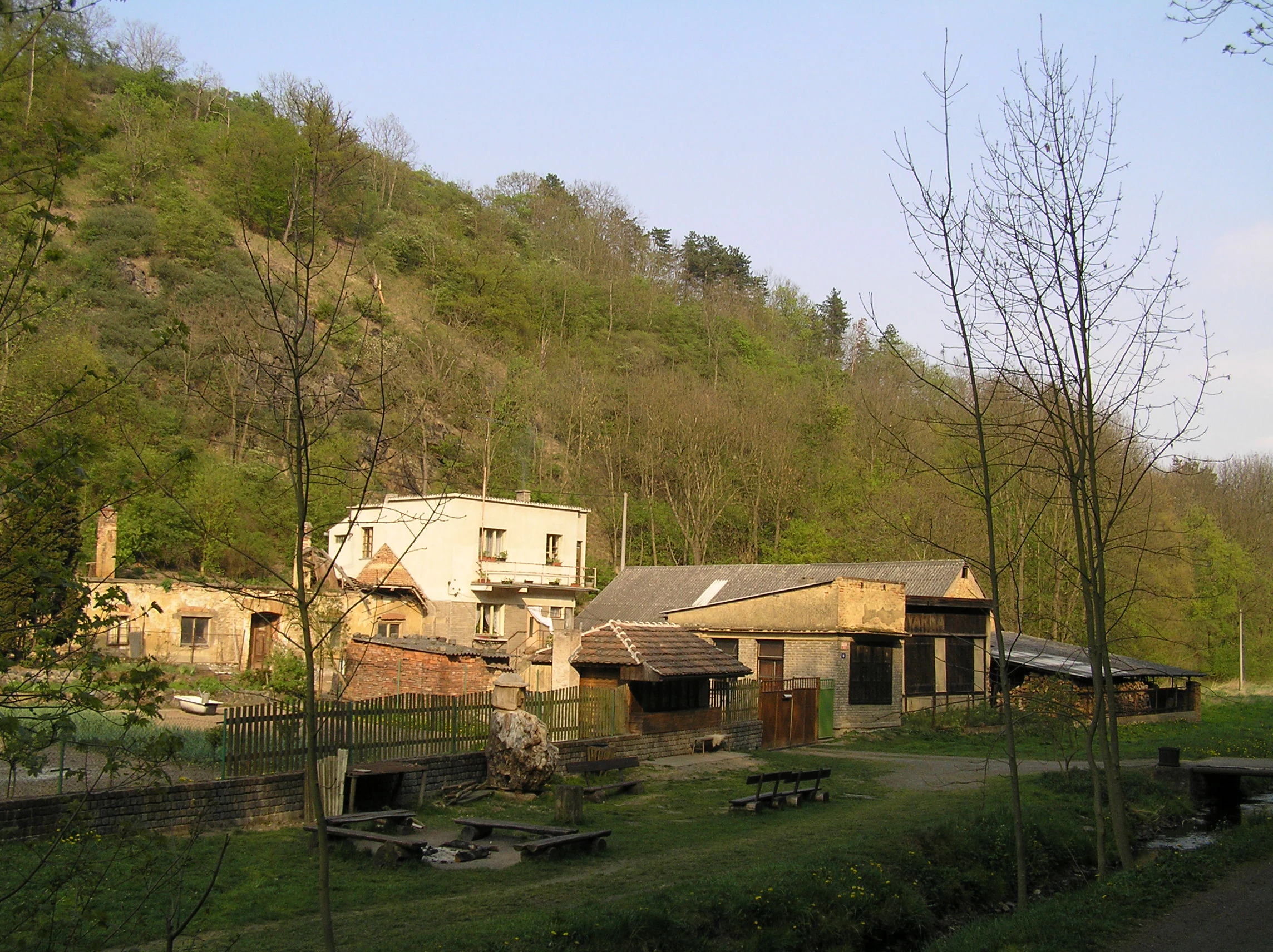 A sloped, forested hillside rising behind a collection of small, older buildings and a grassy area with outdoor seating in a rural setting.