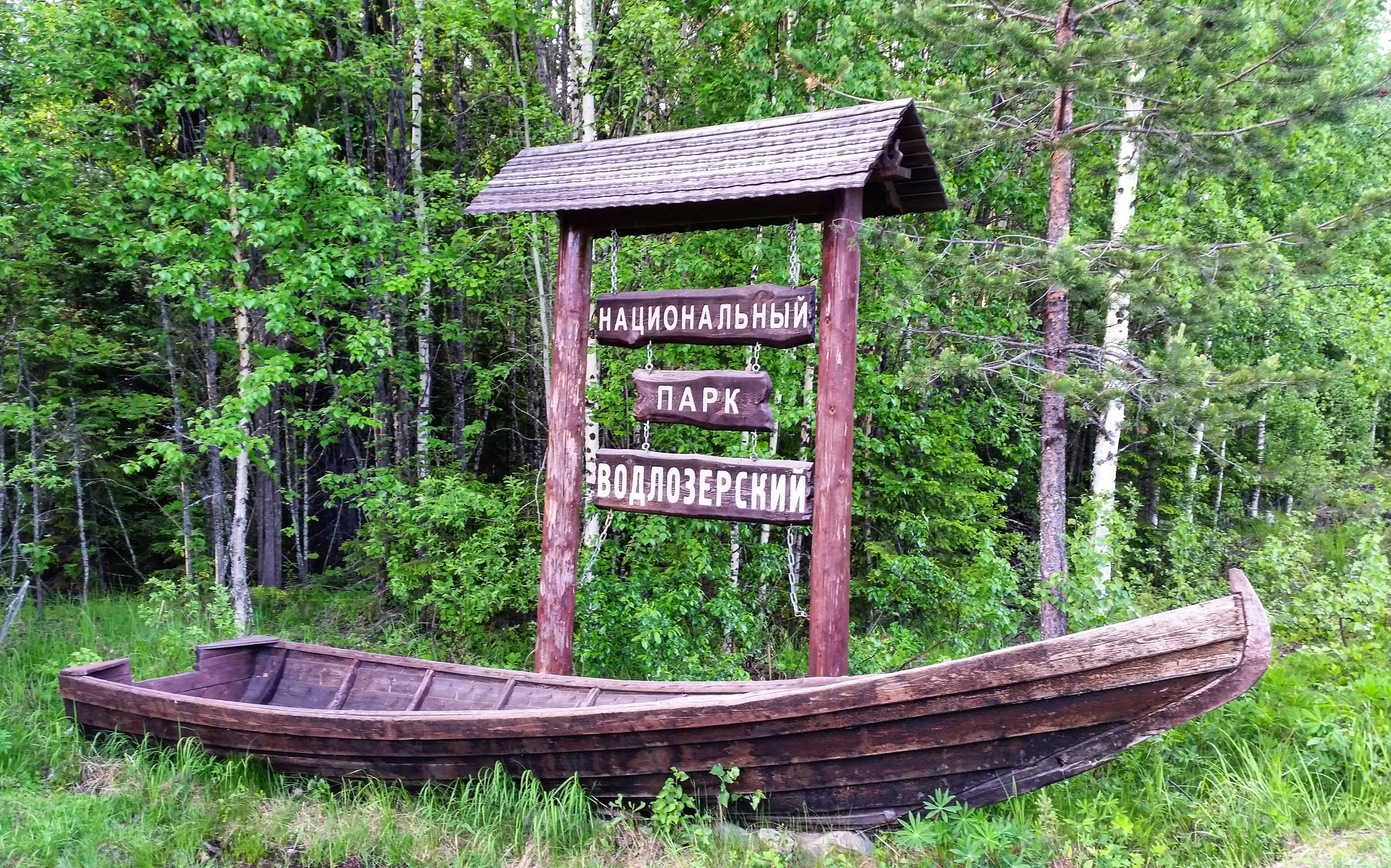 A wooden sign for Vodlozero National Park, Russia, mounted on two vertical logs with a small pitched roof, positioned behind a decorative wooden boat on the ground in a forest setting.