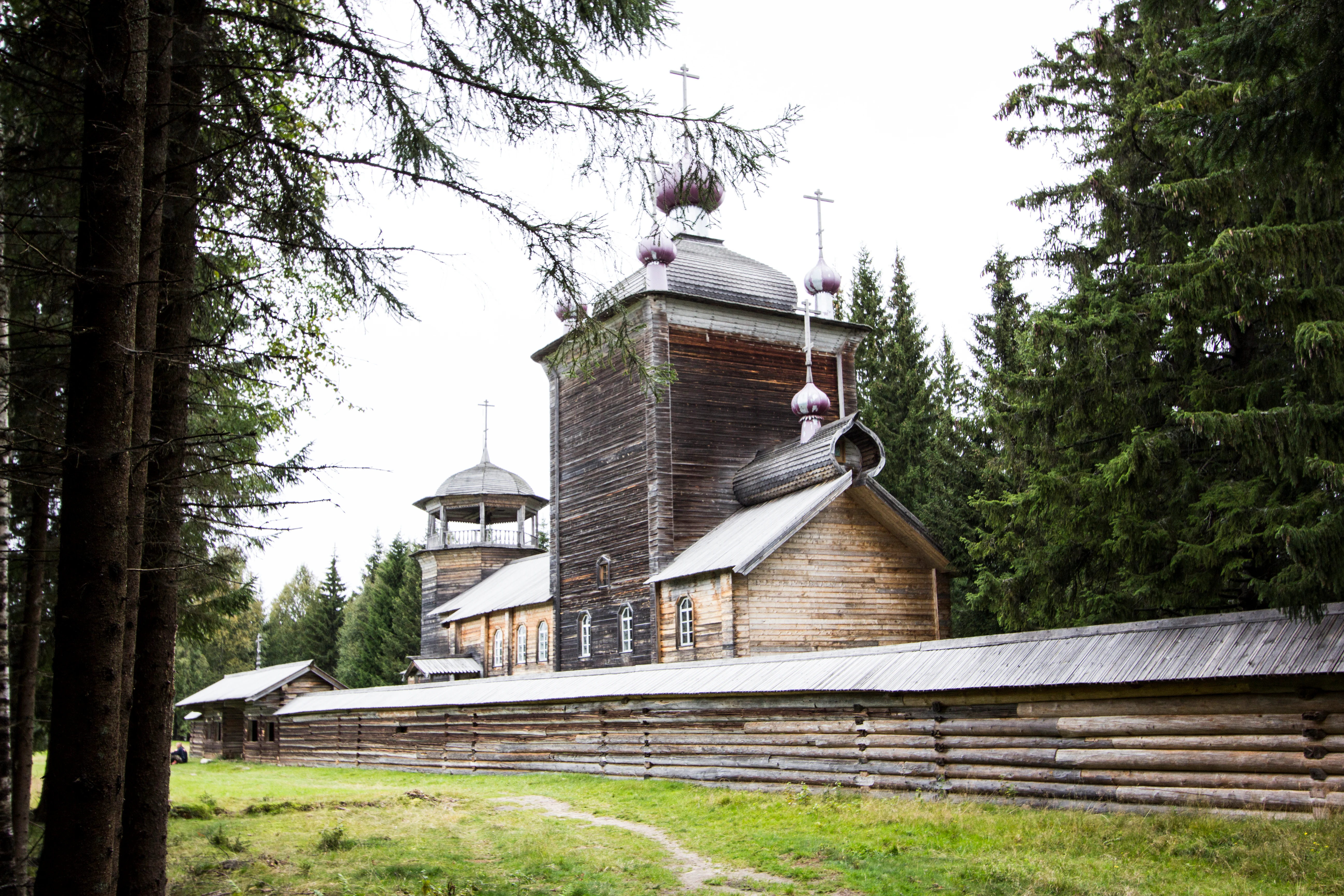 A traditional Russian wooden church with multiple onion domes surrounded by dense coniferous forest and an enclosed log fence.