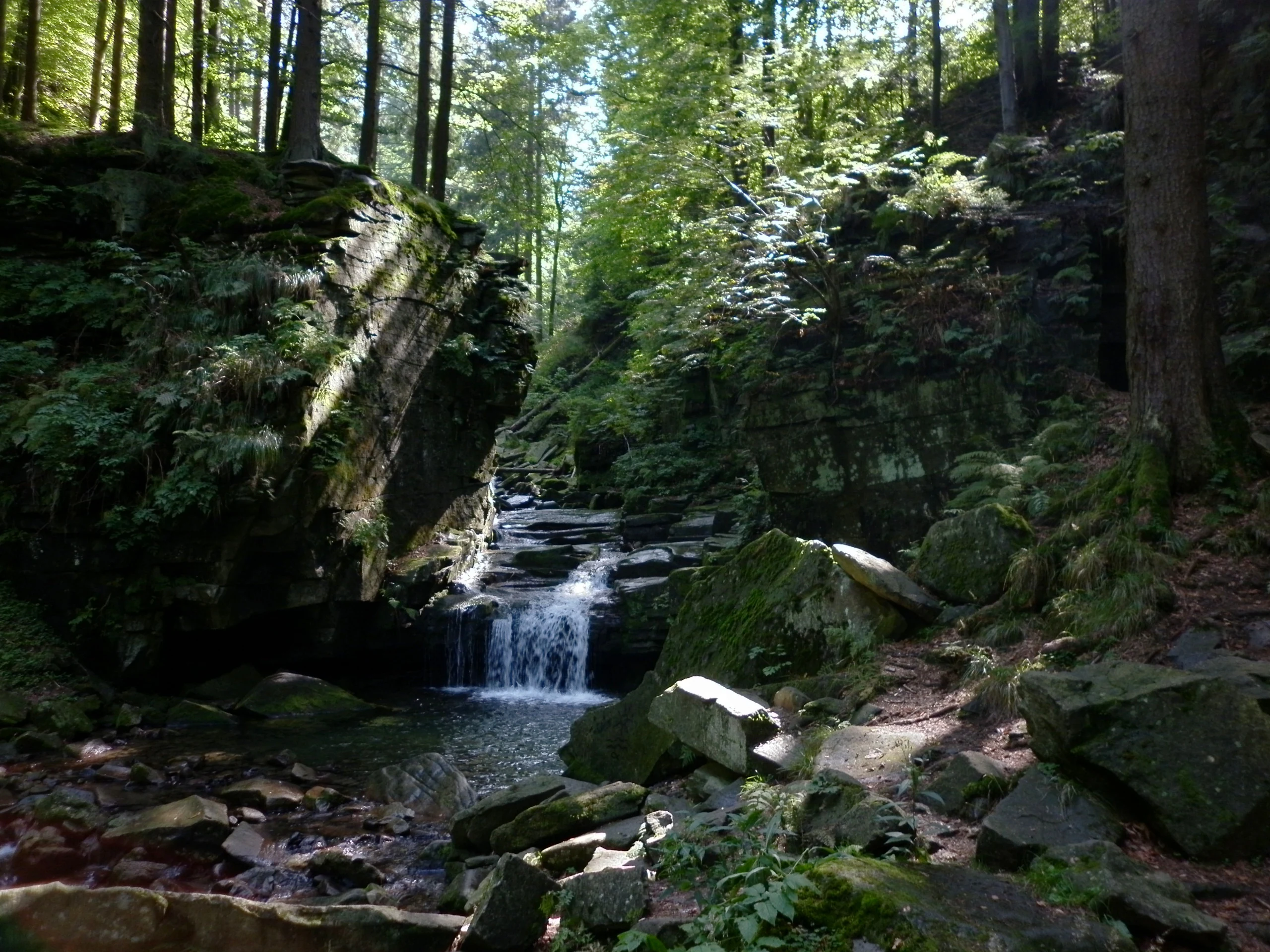 A small waterfall flows over dark rocks into a pool surrounded by a dense forest at the Vodopády Satiny natural monument.
