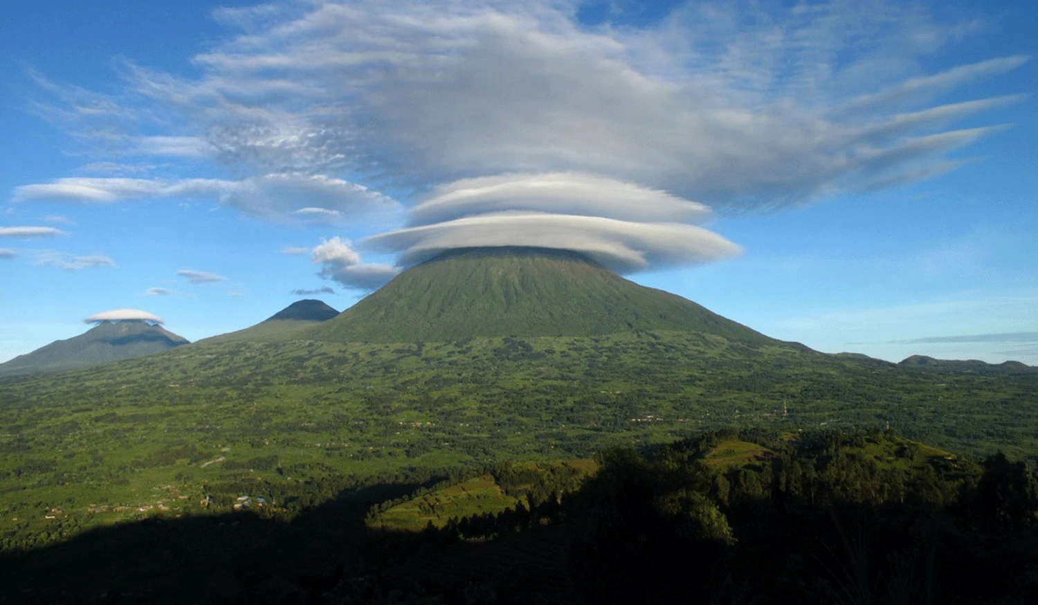 Una montaña con una nube lenticular sobre la cumbre, rodeada de paisaje verde bajo un cielo azul despejado