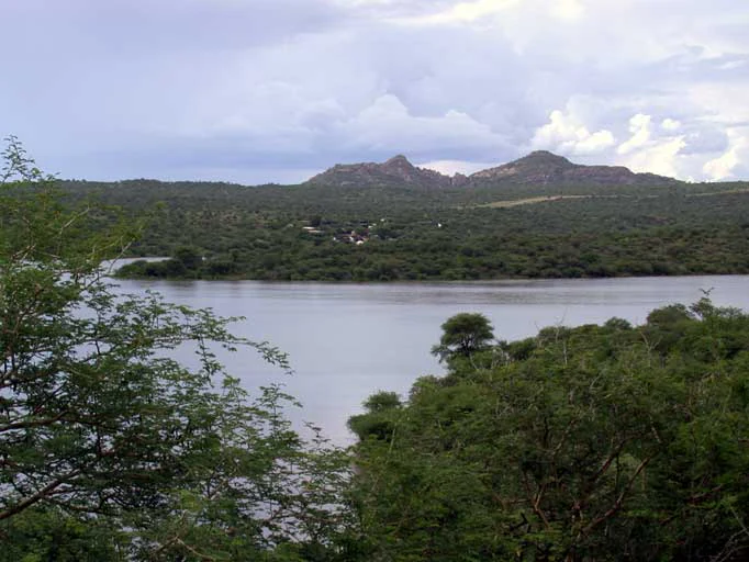 A wide view of the Von Bach Dam reservoir under an overcast sky, with dense green vegetation in the foreground and rocky hills in the distance.
