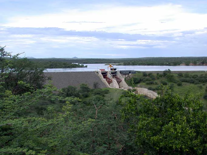 A wide shot showing the Von Bach Dam structure holding back a large reservoir, surrounded by lush green bushveld vegetation under an overcast sky.