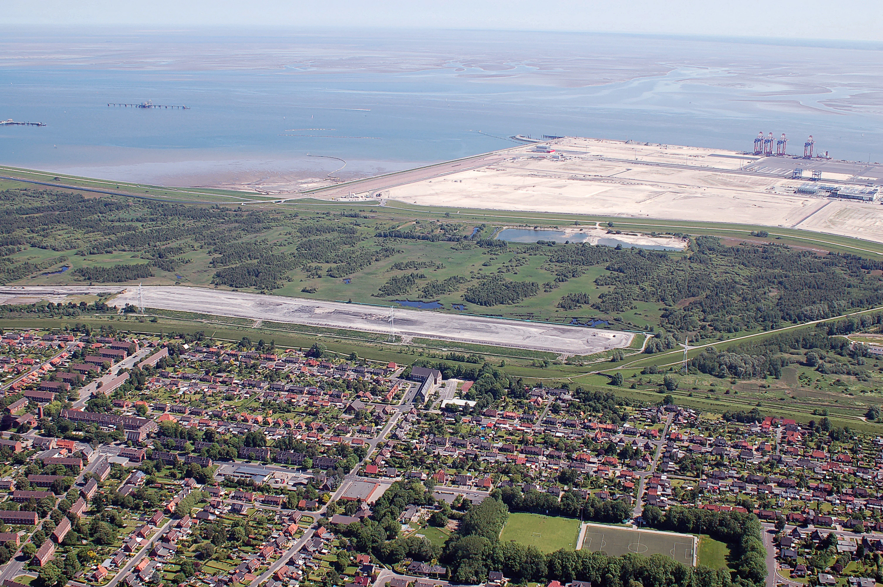 An aerial photograph showing the green, vegetated Voslapper Groden-Süd nature reserve situated between a residential district in the foreground and the North Sea coastline with large industrial facilities in the background.