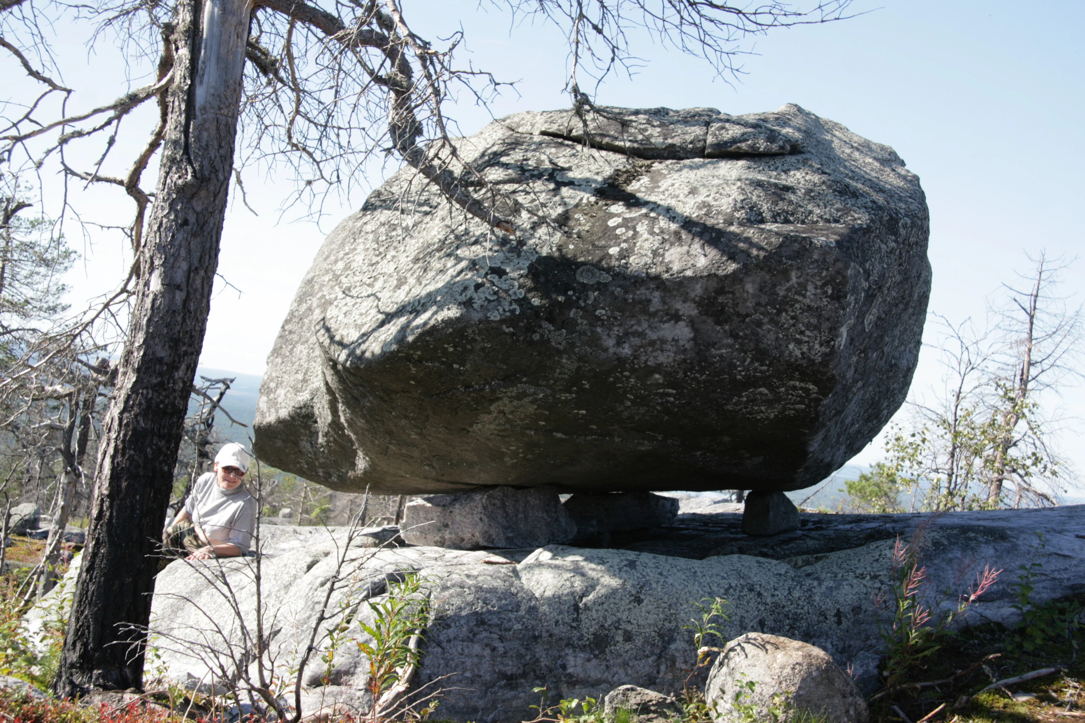 A large, rounded grey boulder balanced on three smaller stones atop a rocky base, with a person sitting nearby for scale and sparse trees in the background.