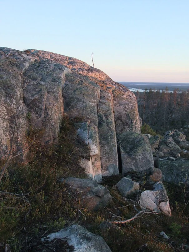 A large, weathered rock face featuring vertical fissures and nearby scattered boulders set against a backdrop of distant forest and a pale horizon at dusk.