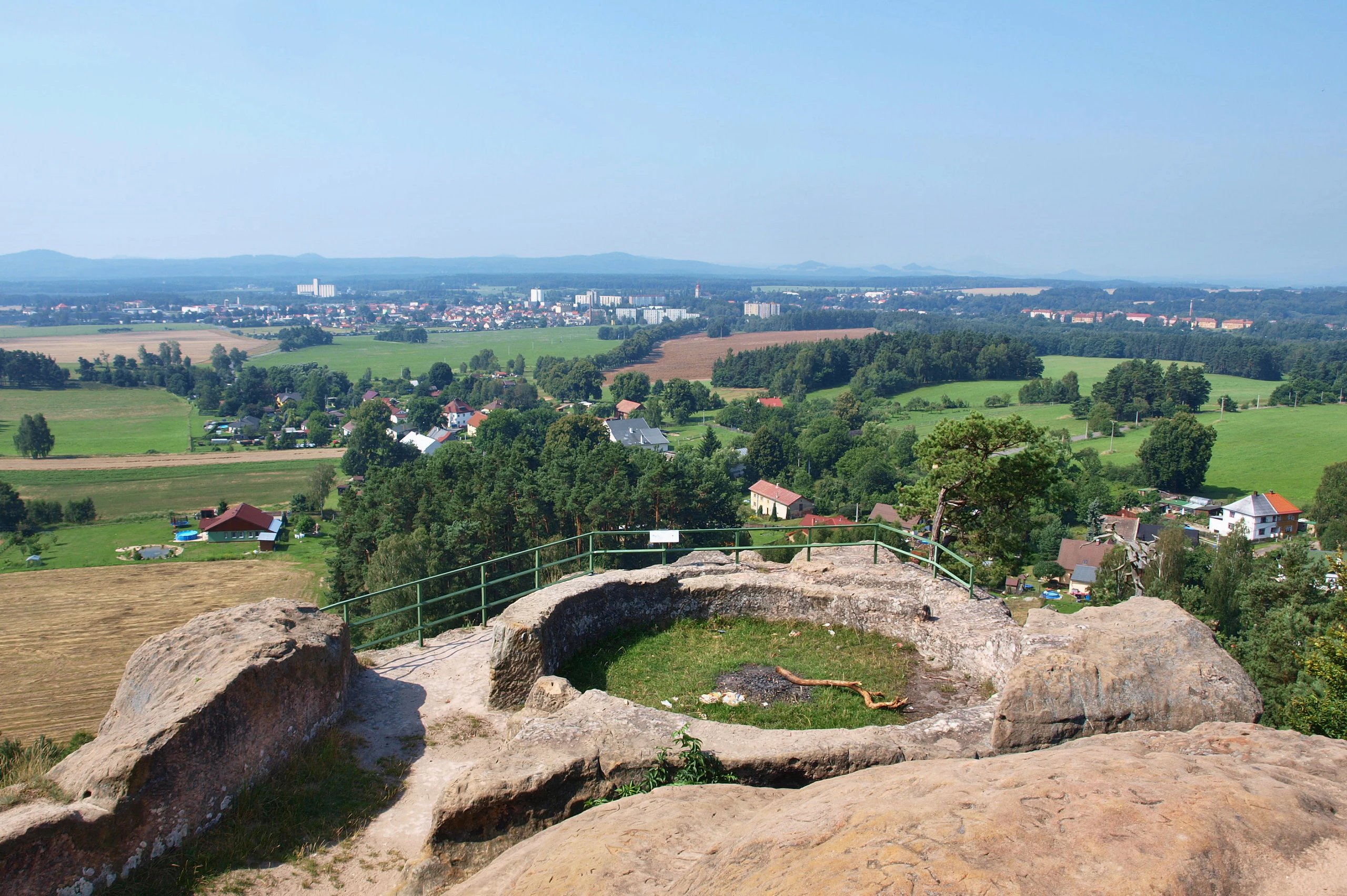 A high angle view from a rocky sandstone overlook with a green metal railing, looking down at a town, green fields, and distant forests under a clear blue sky.