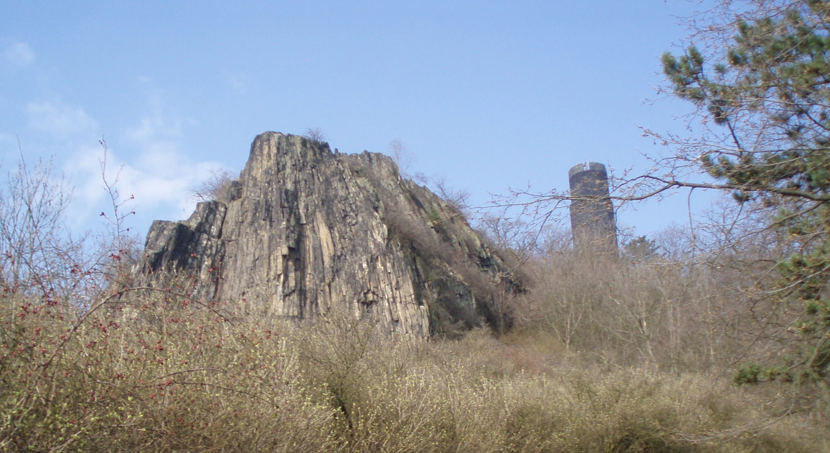 A large basalt rock formation and a stone tower rising above dry, grassy slopes under a clear blue sky.