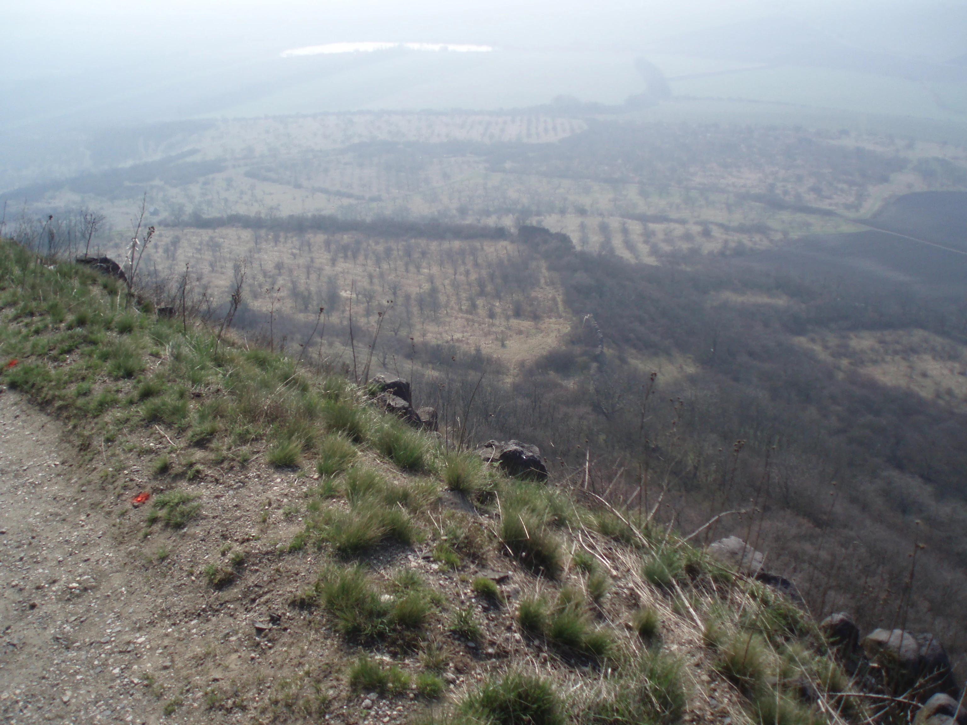 A high-angle view looking down from a grassy, rocky hillside trail across a hazy landscape of fields and sparse orchards.
