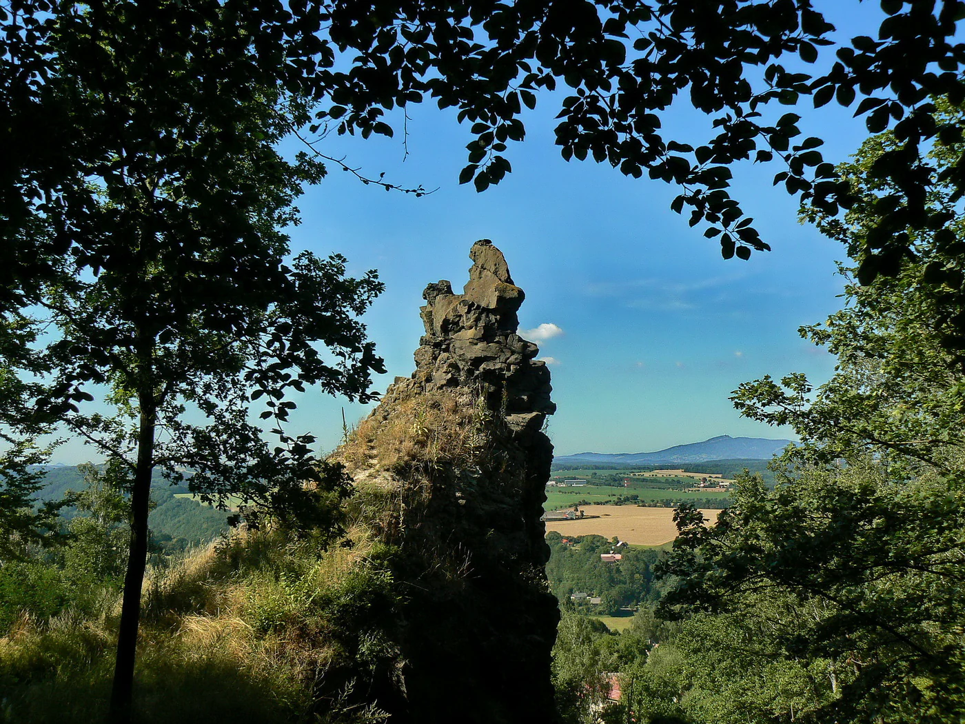 A prominent, jagged rock formation stands in the center foreground, surrounded by trees. In the background, rolling fields and a distant mountain peak are visible under a clear blue sky.