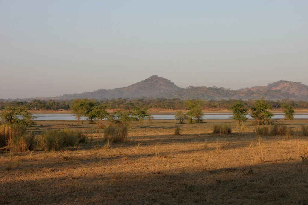 A wide, golden-hour view of Lake Kazuni, showing a calm body of water flanked by sparse trees and dry grasses, with rolling hills in the background.
