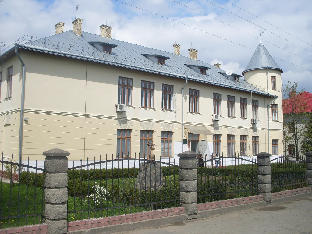 Beige two-story building with gray metal roof and multiple chimneys, surrounded by black metal fence with stone pillars, green landscaping, and clear sky