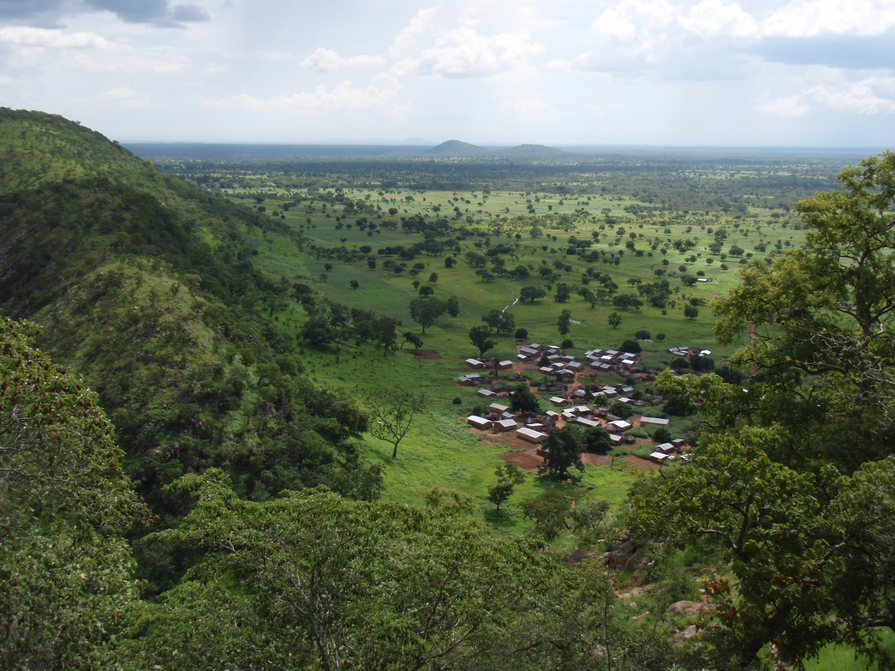 A high-angle view looking down over a lush, green valley filled with scattered trees, with a small village visible in the midground and the Atakora mountain range rising on the left.