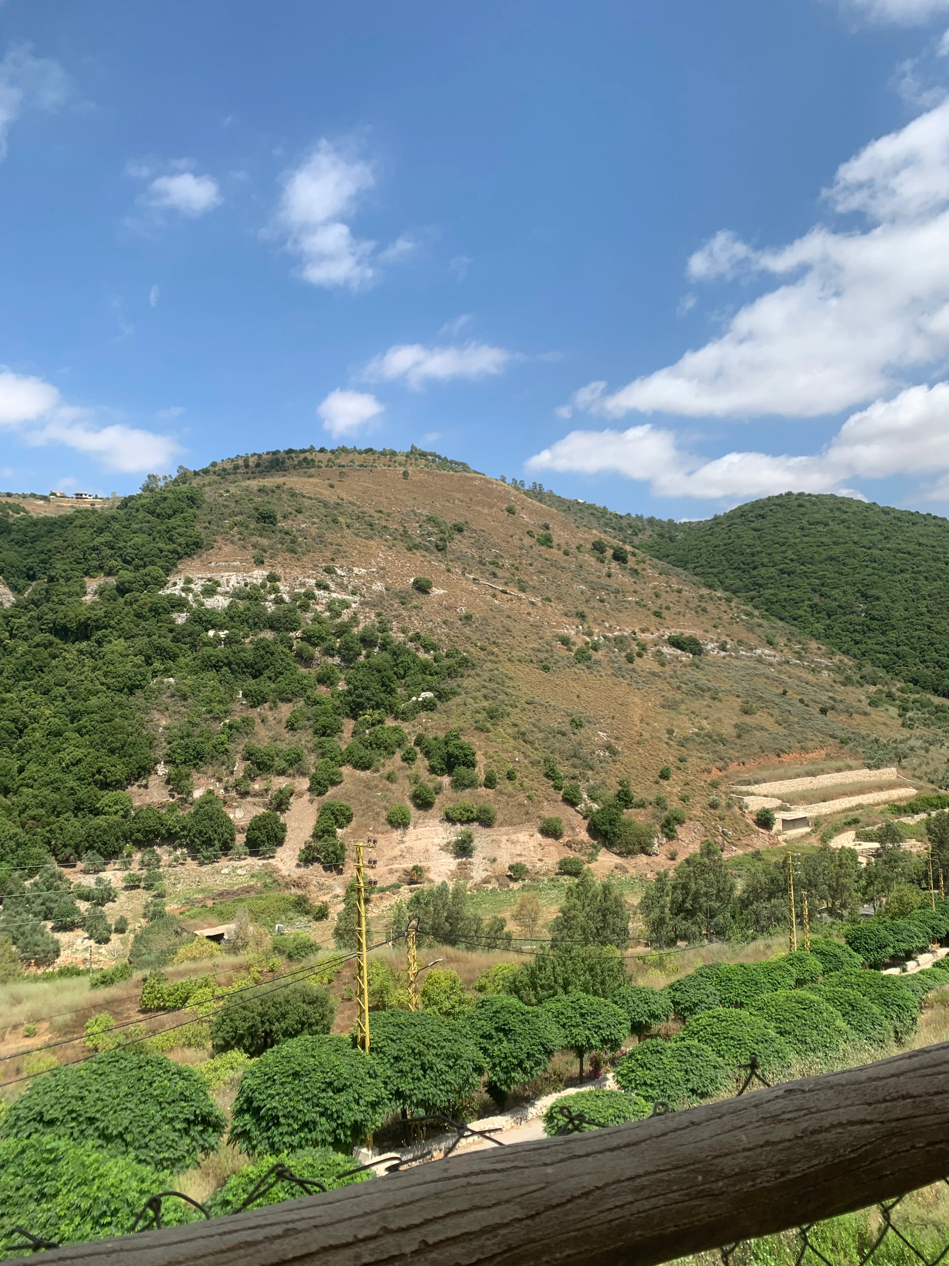 Una toma amplia que mira a través de un valle hacia colinas onduladas cubiertas de arbustos, árboles y secciones agrícolas en terrazas bajo un cielo azul brillante.