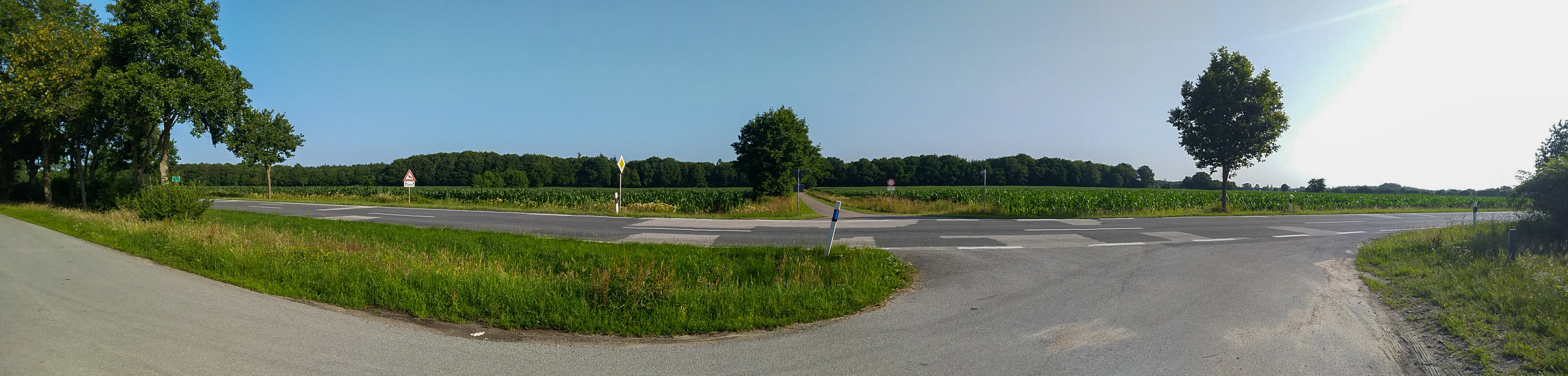 A wide panoramic shot of a rural road intersecting with an agricultural field and a dense treeline in the background under a clear sky.