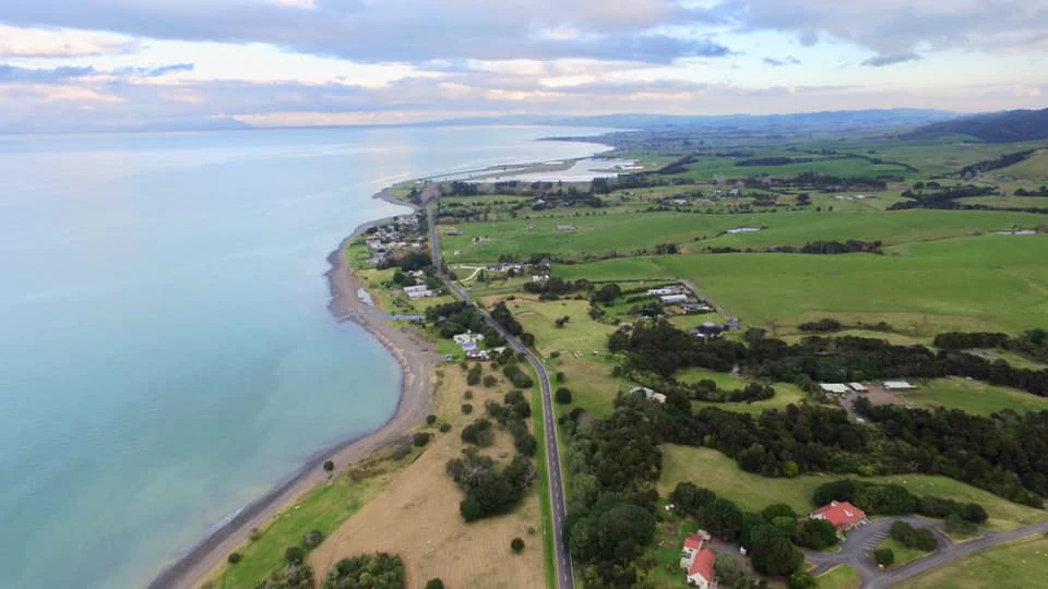 An aerial view of a road running parallel to a coastline with a large body of water on the left, flanked by green fields and scattered trees on the right.