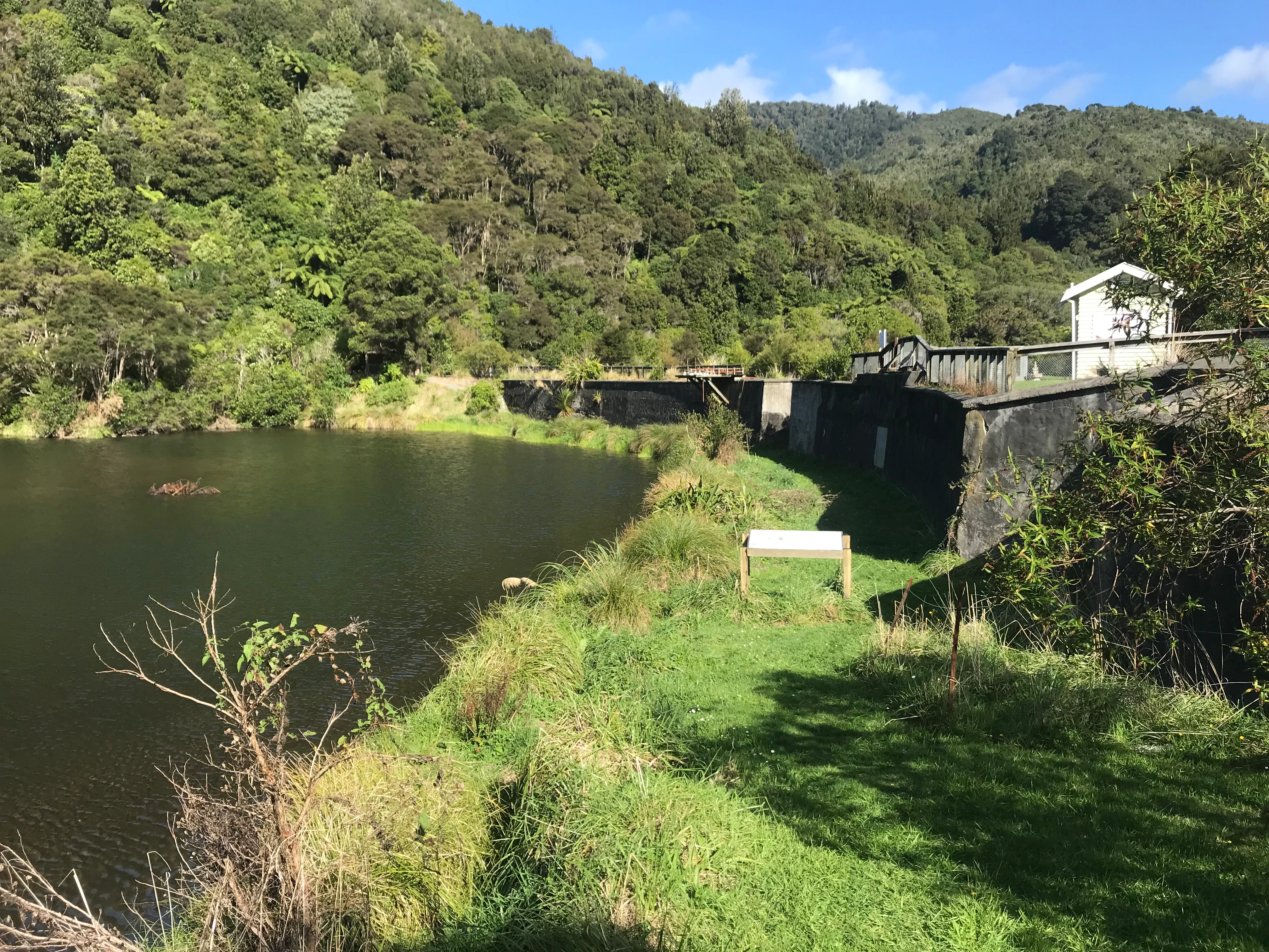 A historic stone dam structure next to a calm reservoir in a lush, green, forested valley.